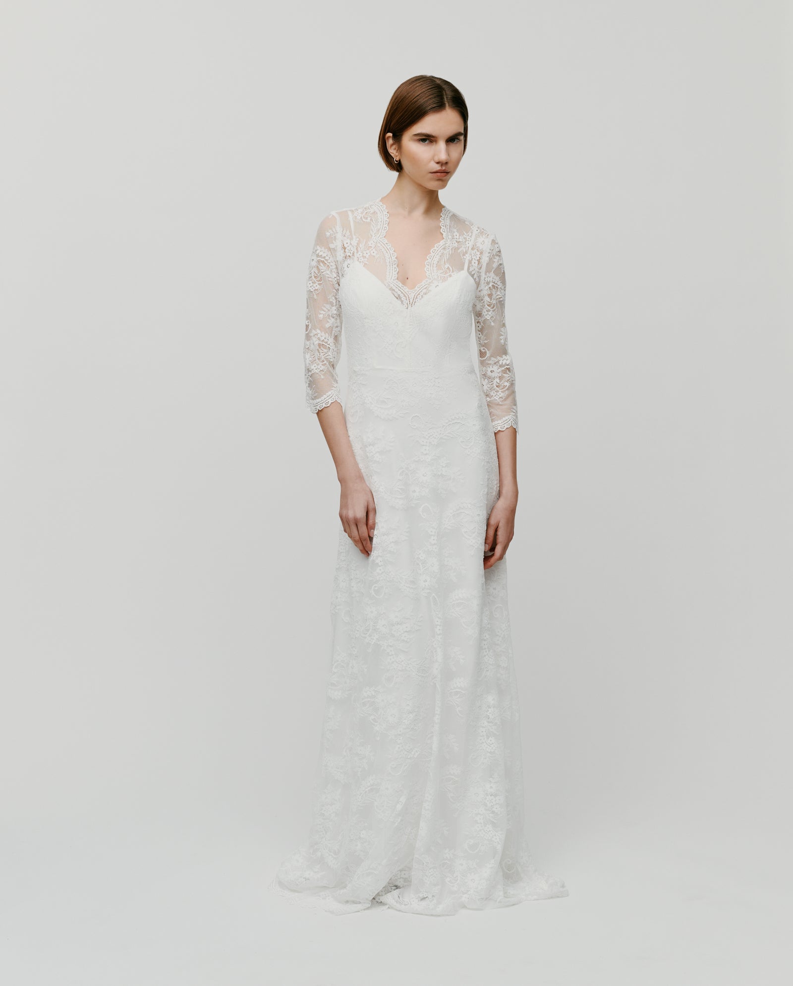 Woman wearing the IVY OAK DALIA AMY Dress, a long white bridal lace gown with a V-neckline and three-quarter sleeves, standing against a plain background.