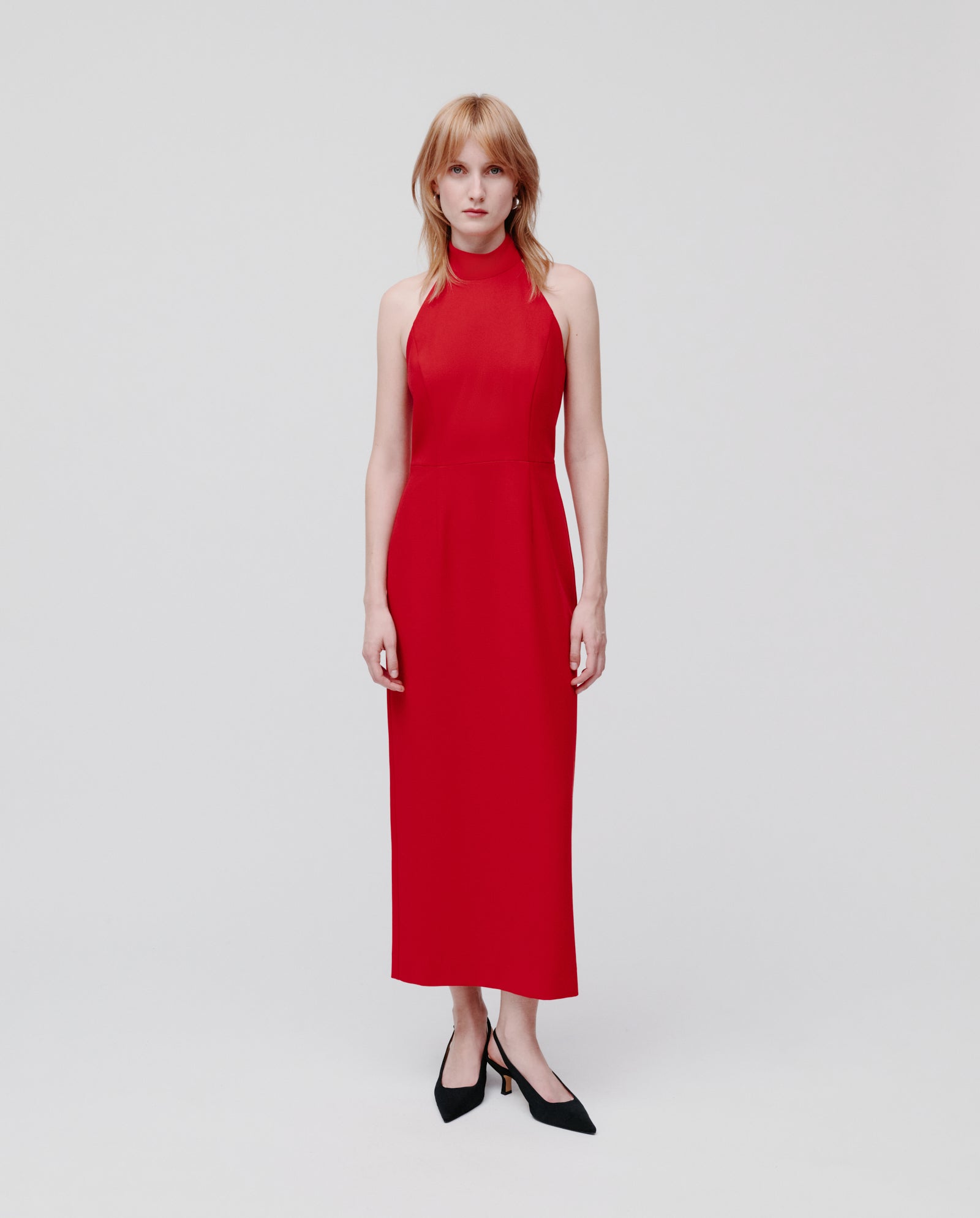 A woman wears the sleeveless, red NOLAN Dress by IVY OAK, featuring a body-hugging fit and neckholder design, paired with black heels, standing against a plain white background.