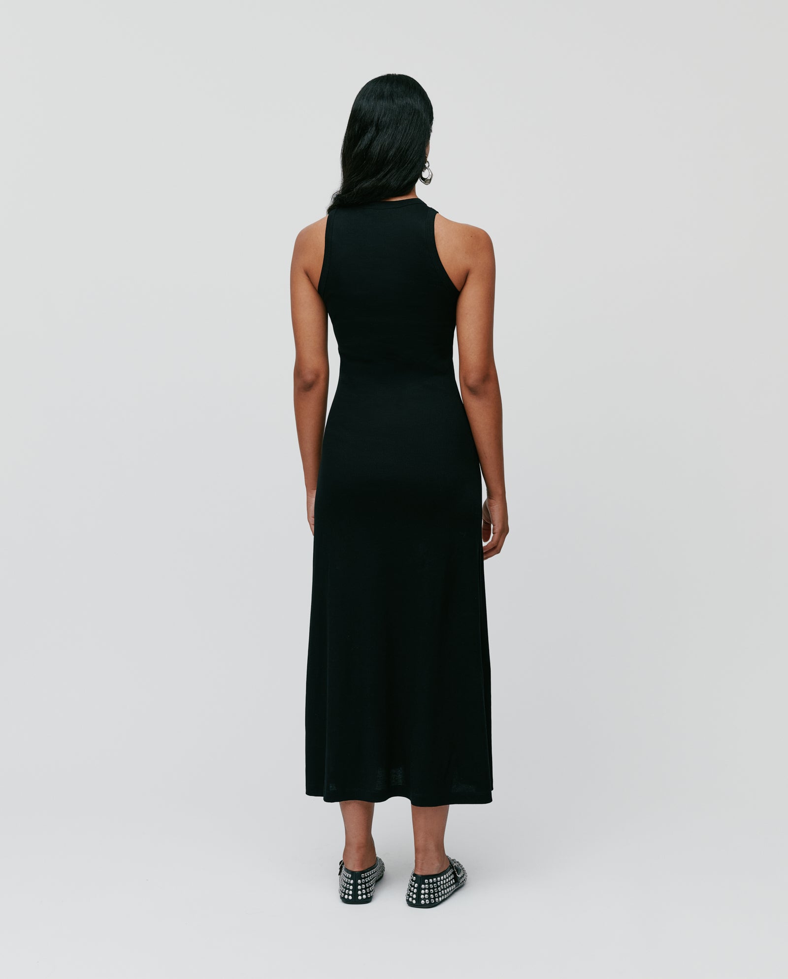 A woman faces away from the camera, wearing the IVY OAK KRISTYNA sleeveless ribbed knit black midi dress with a flared skirt and studded shoes.