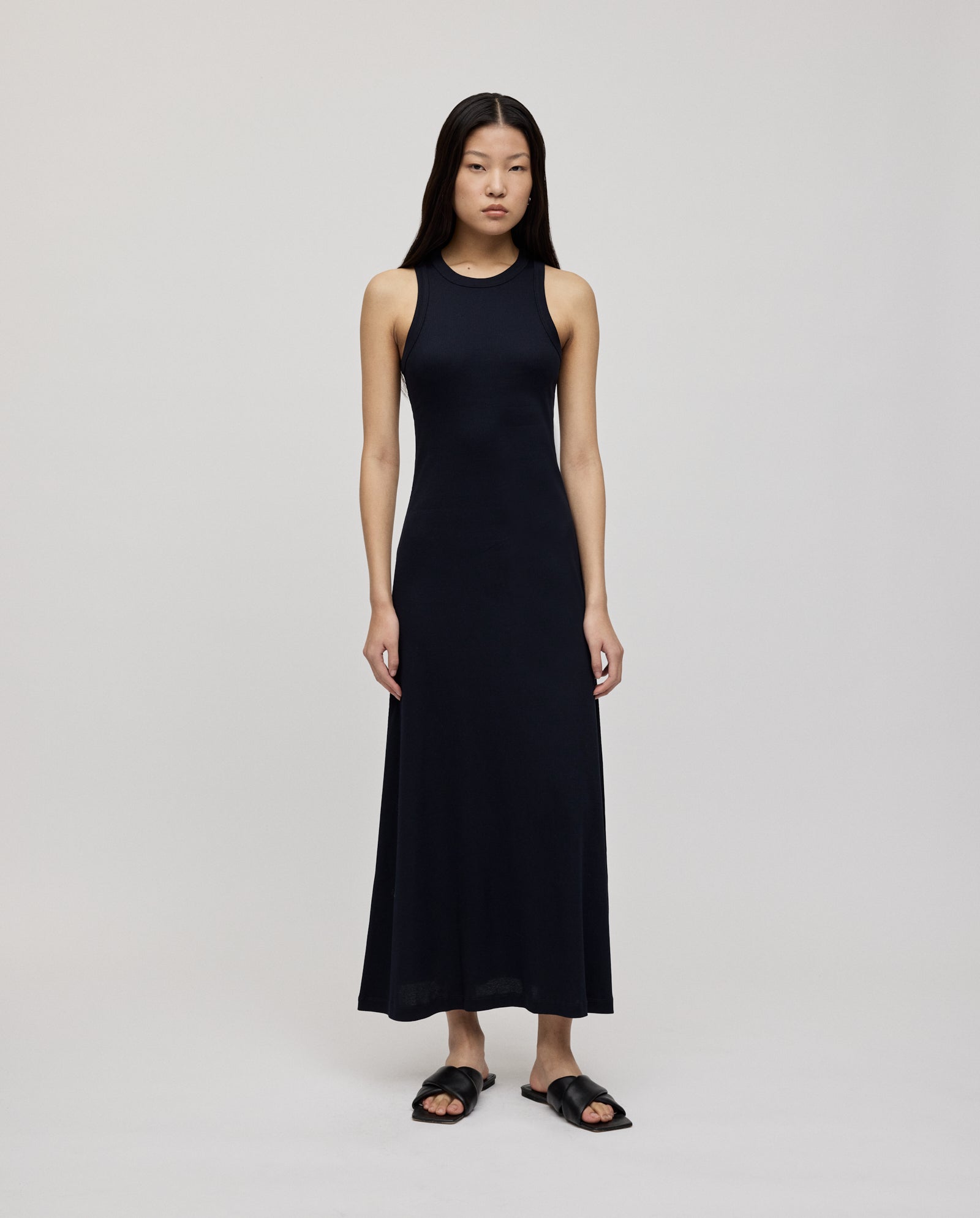 A woman wears the IVY OAK KRISTYNA sleeveless long black ribbed knit dress with a flared skirt and black sandals, standing against a plain light background.