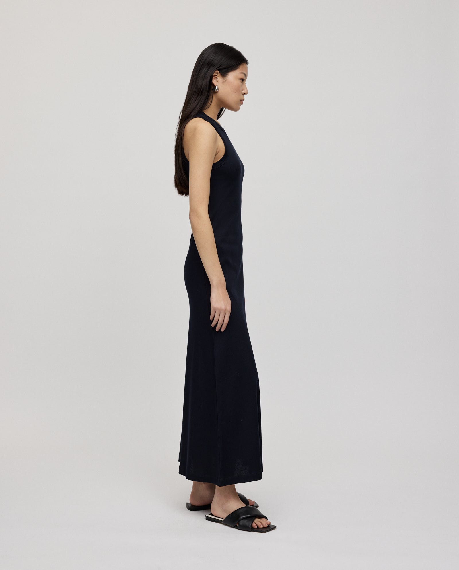 A woman in a sleeveless black IVY OAK KRISTYNA Dress with a flared skirt and black sandals stands in profile against a plain background.