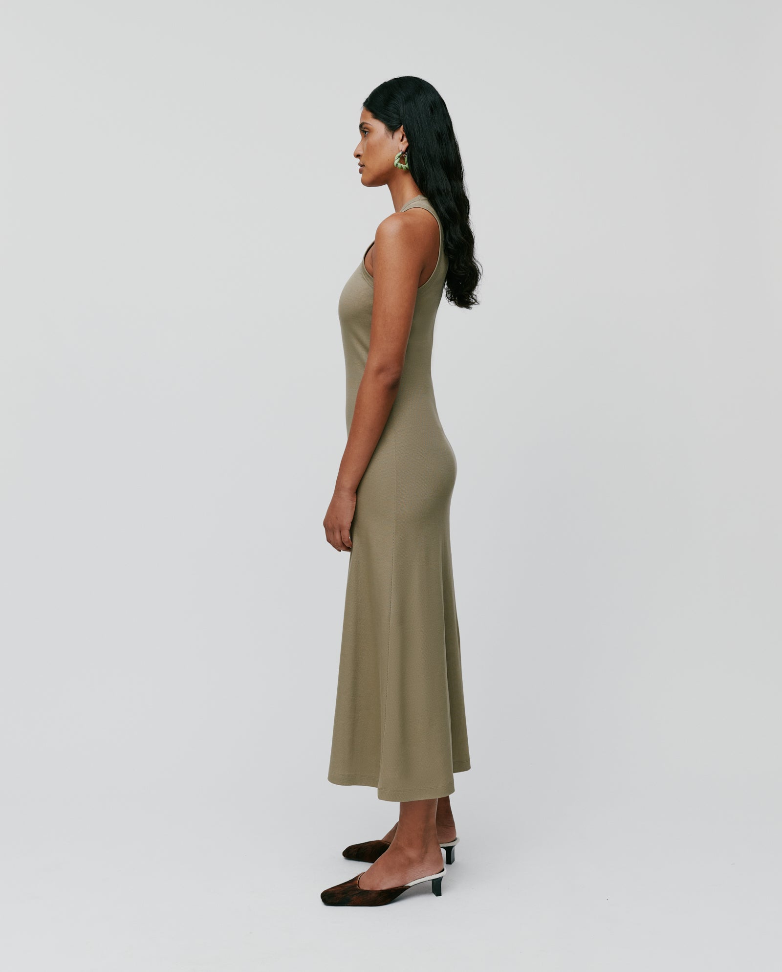 A woman in the KRISTYNA sleeveless olive green dress by IVY OAK, featuring a subtle flared skirt, paired with black heels, stands in profile against a plain light background.