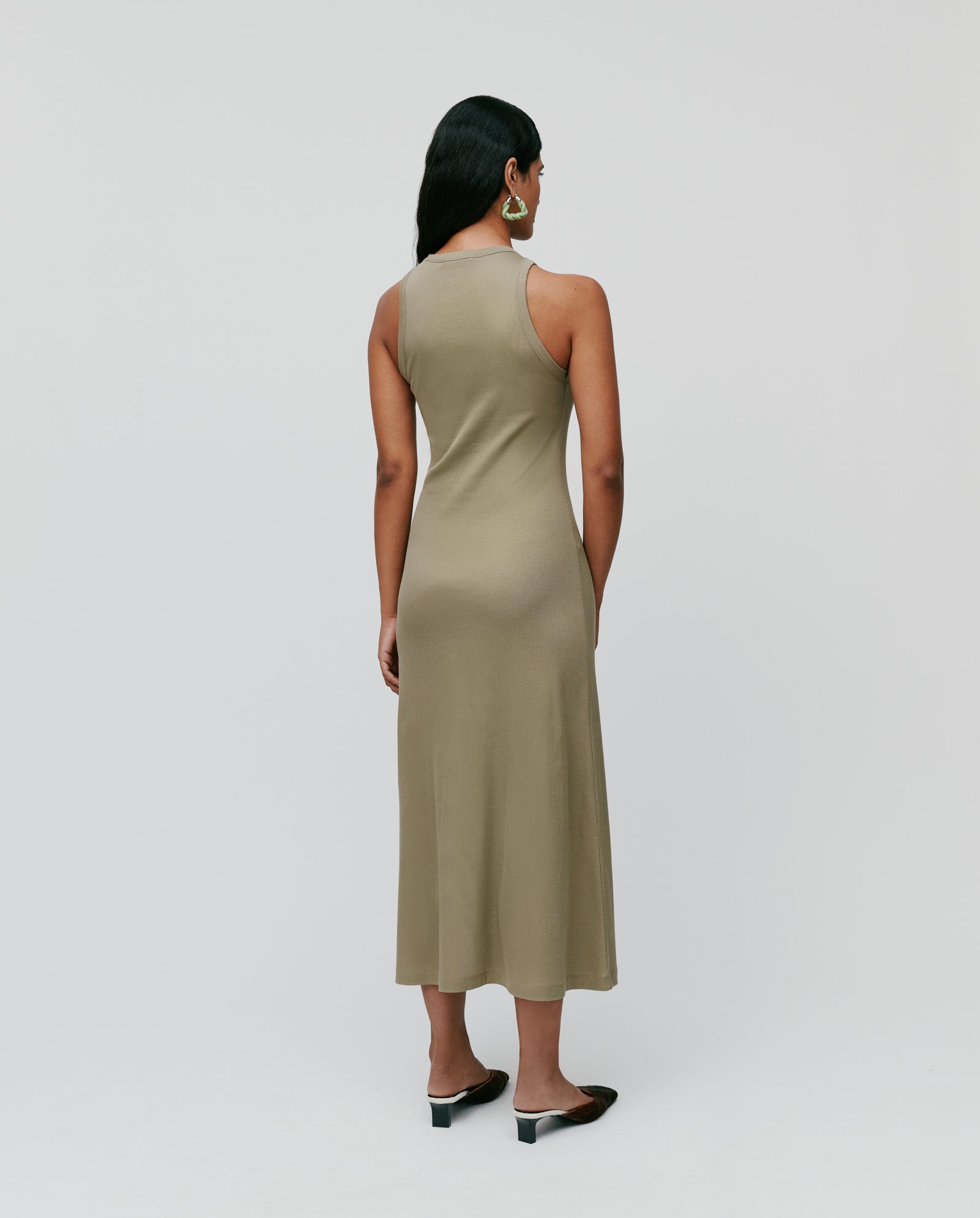 A woman stands in profile against a plain background, wearing the IVY OAK KRISTYNA Dress—an olive-green, sleeveless midi with a flared skirt—paired with black heels.
