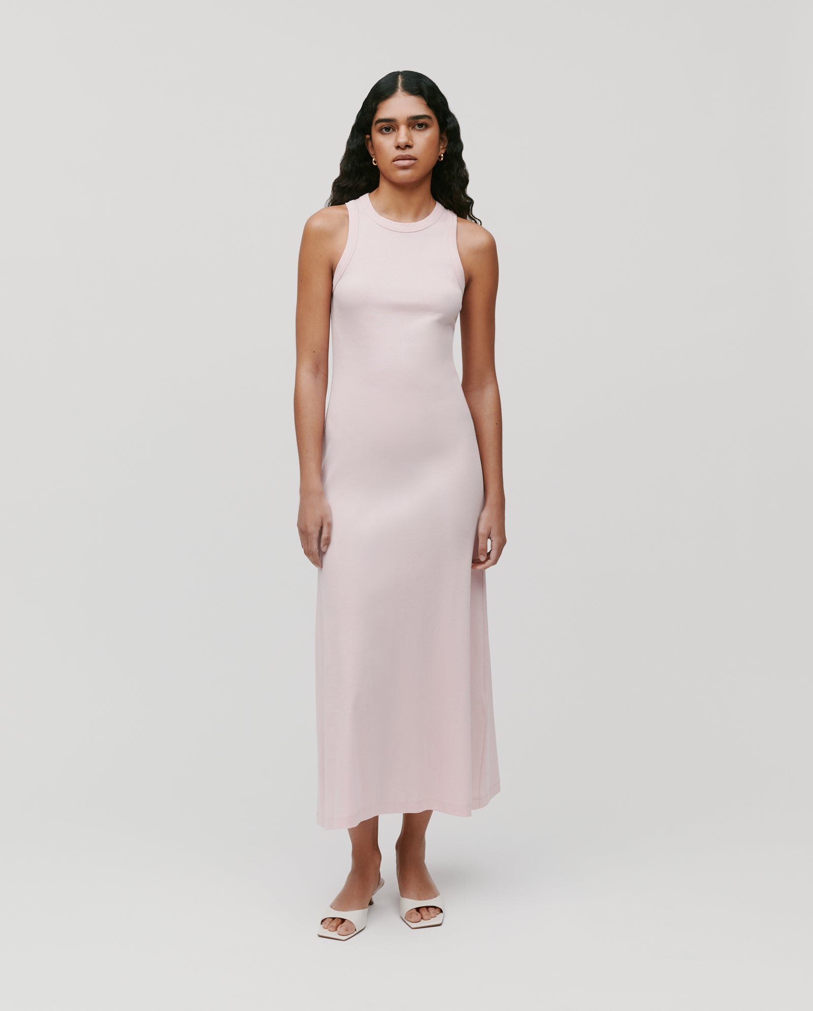 A woman wears the KRISTYNA Dress by IVY OAK, a sleeveless light pink dress with a flared skirt, paired with white sandals and posed against a plain background.