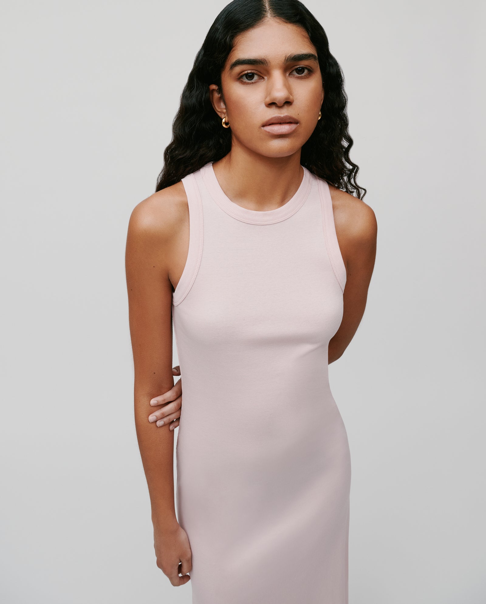 A woman with long wavy hair wears the KRISTYNA sleeveless light pink ribbed knit dress by IVY OAK, standing against a plain background.