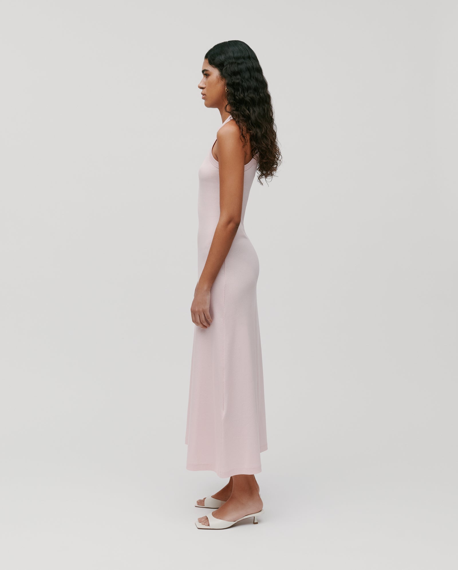 A woman in profile wears the IVY OAK KRISTYNA Dress, a light pink sleeveless style with a flared skirt, paired with white sandals, set against a plain white background.
