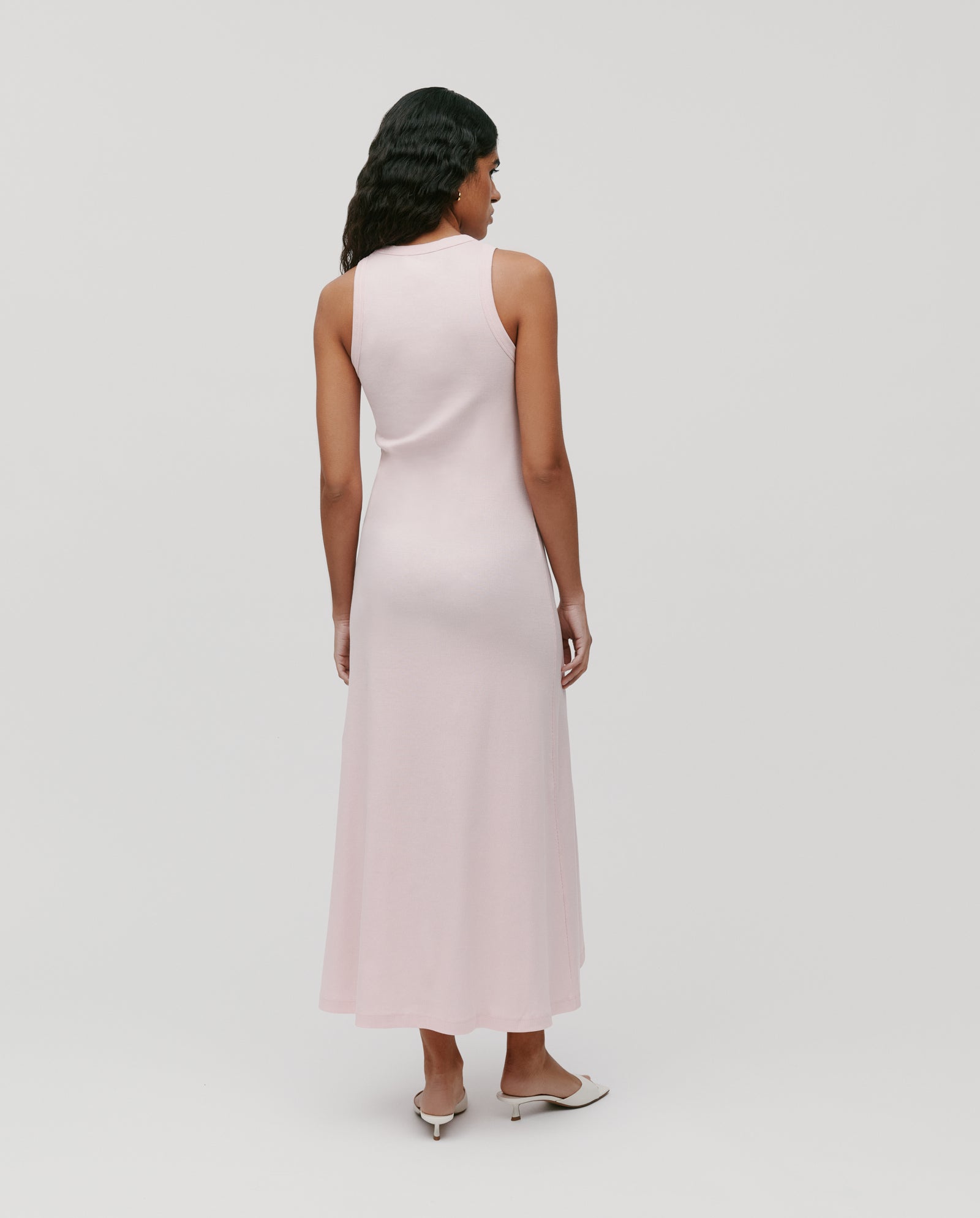 A woman stands with her back to the camera wearing the IVY OAK KRISTYNA sleeveless light pink ribbed knit maxi dress with a flared skirt, paired with white heels.