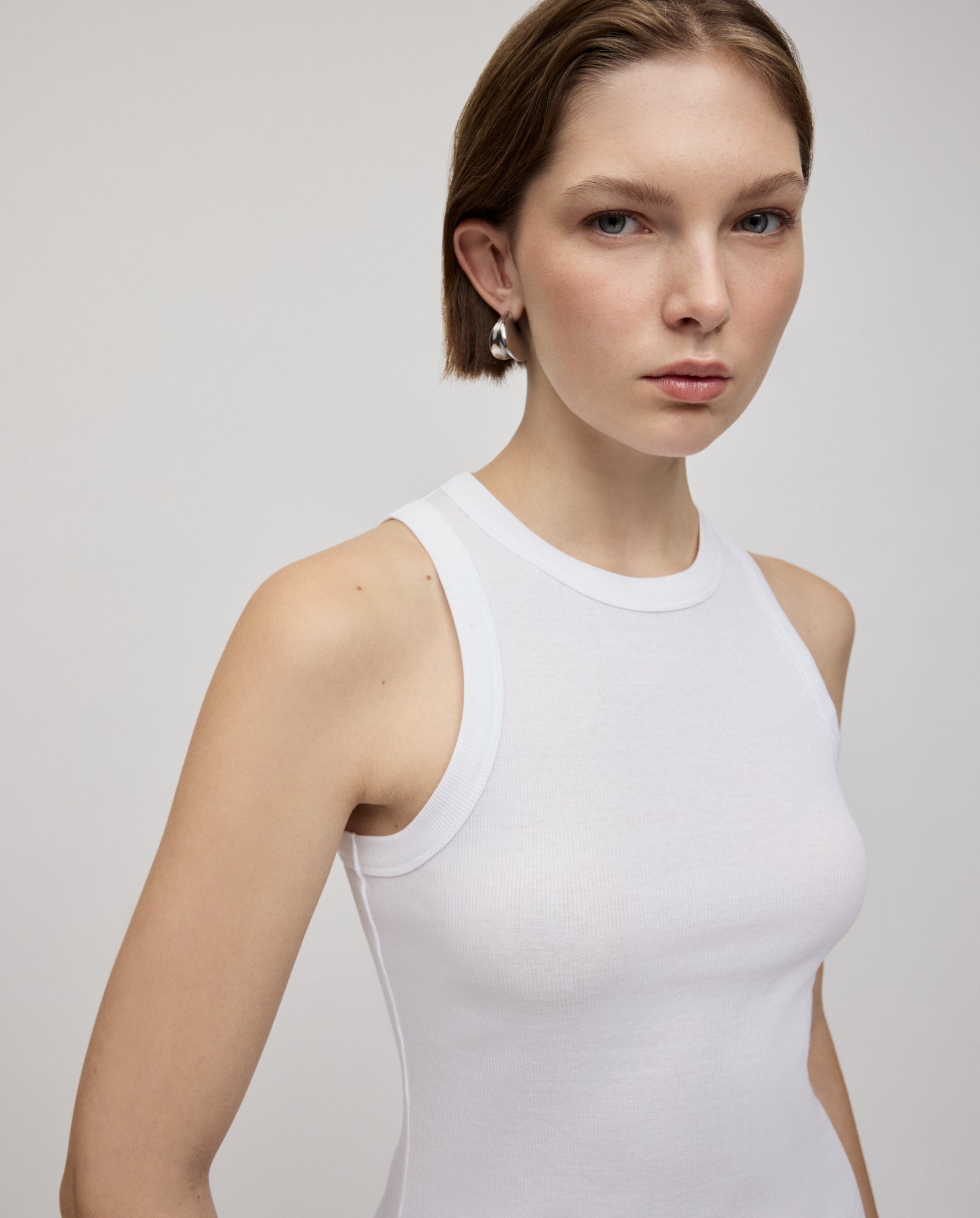 A woman with short hair wears the sleeveless white KRISTYNA Dress by IVY OAK, styled with silver earrings and looking at the camera for a modern look.