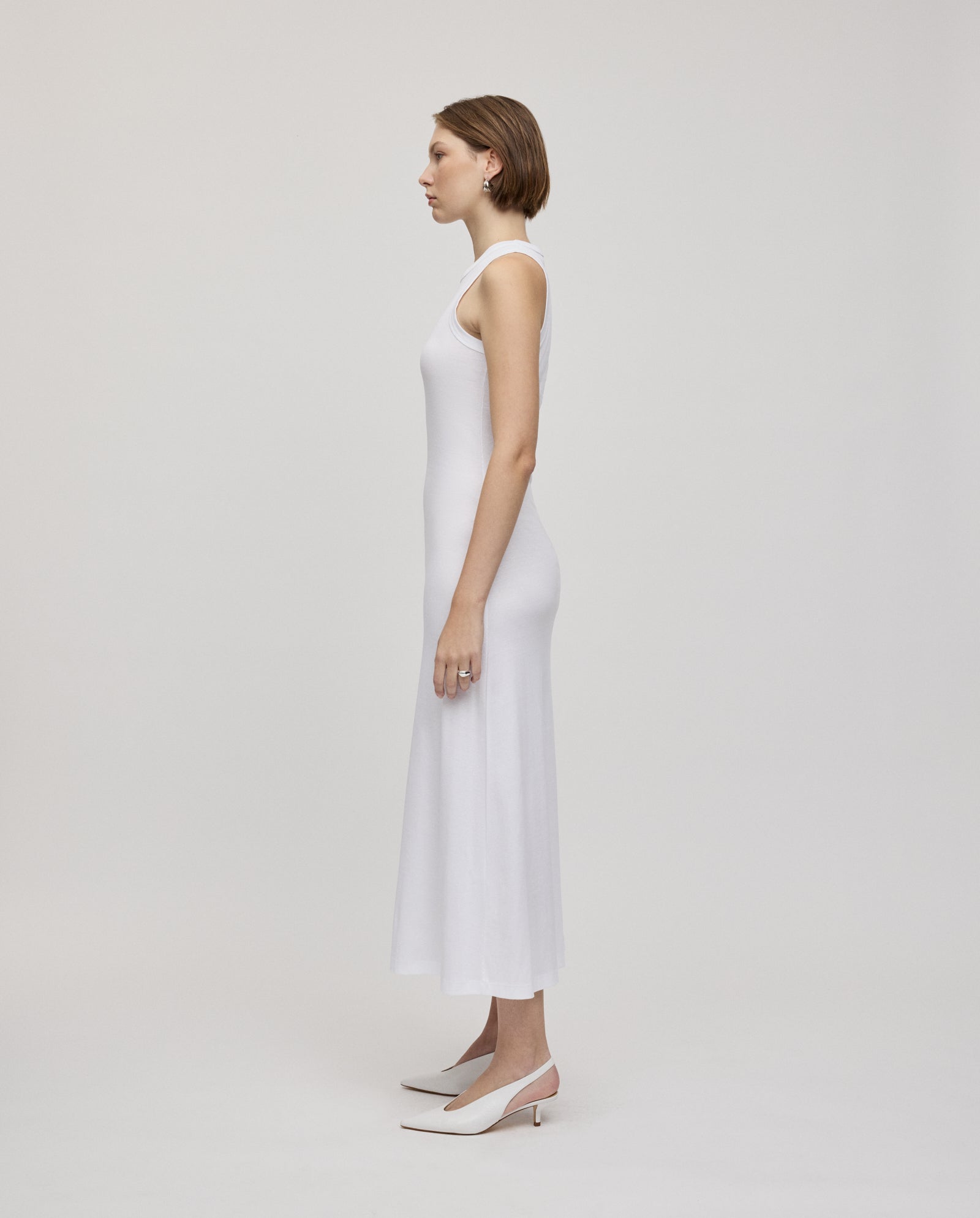 A woman in a sleeveless white IVY OAK KRISTYNA Dress with a flared skirt and white heels stands in profile against a plain white background.