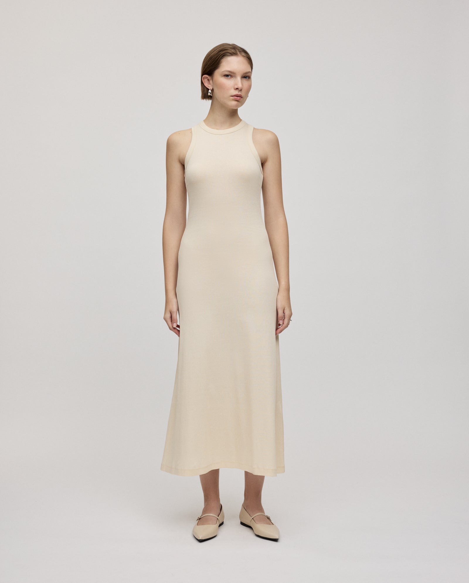 A woman in the IVY OAK KRISTYNA sleeveless, light beige ribbed knit maxi dress with a flared skirt and flats stands against a plain light background.