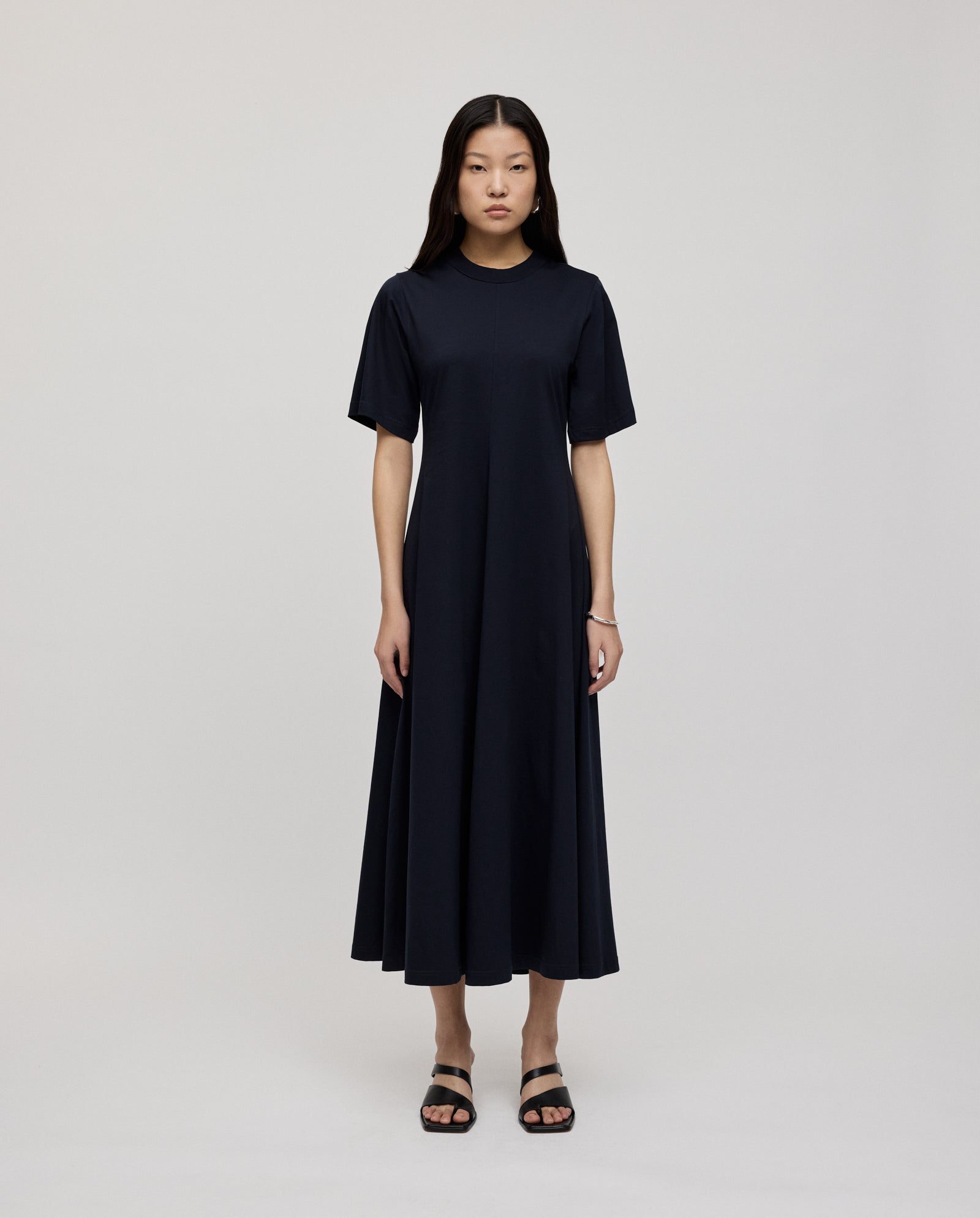 A woman stands against a white background wearing the IVY OAK FRANZISKA Jersey Dress in plain black with a fitted silhouette, paired with black sandals.