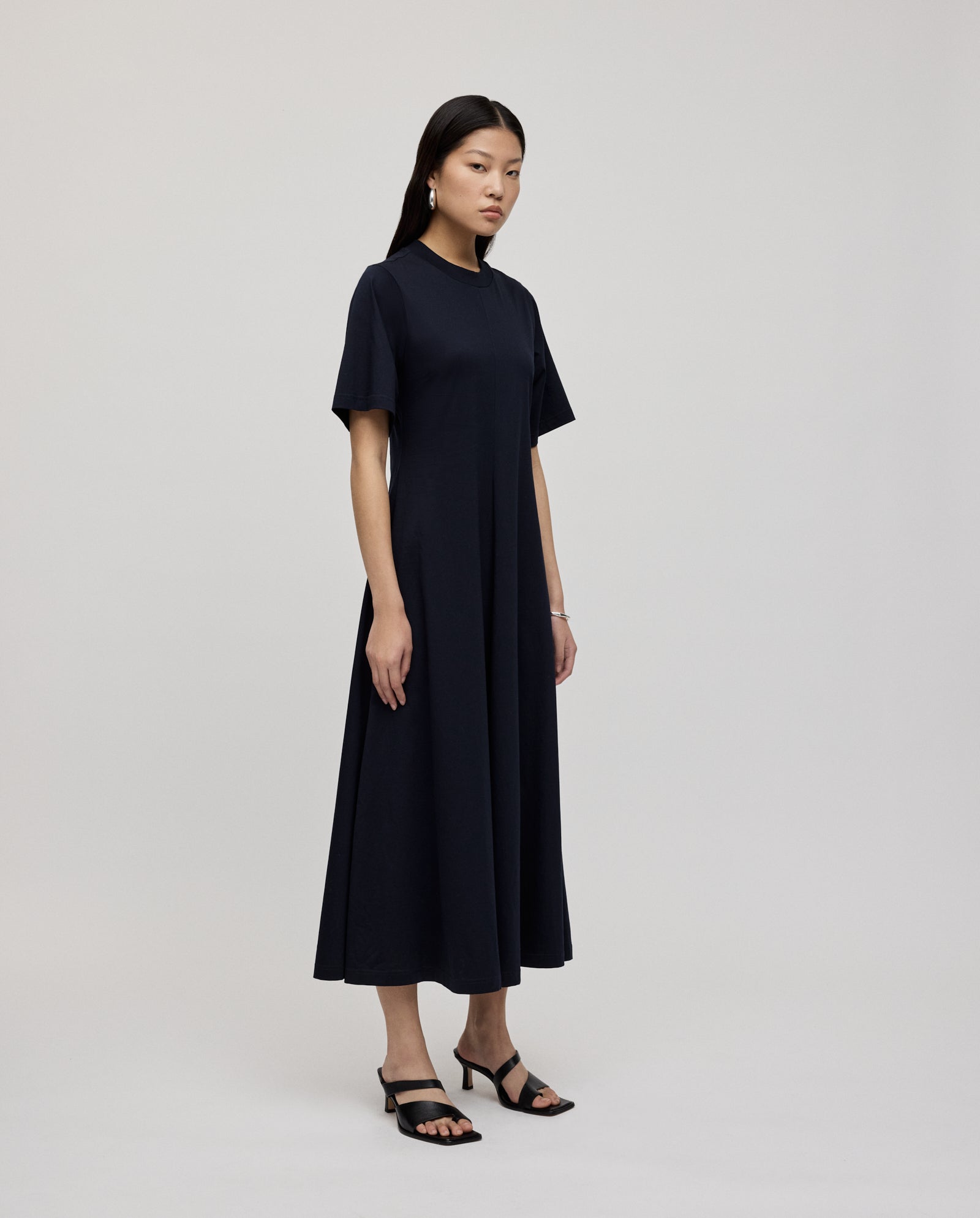 A woman in a black IVY OAK FRANZISKA Jersey Dress with a fitted silhouette and black sandals stands against a plain background.