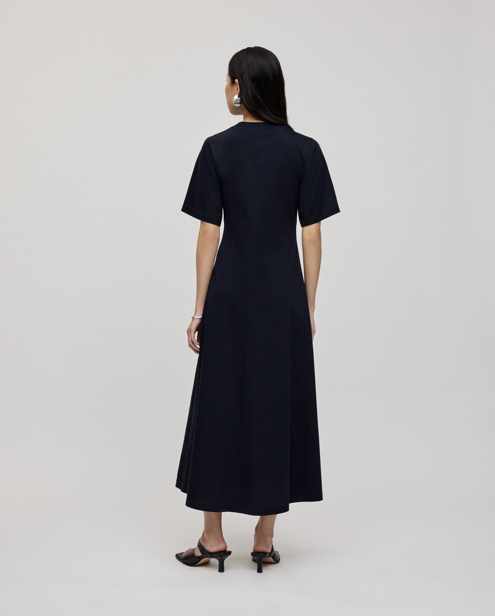 A woman models the IVY OAK FRANZISKA Jersey Dress in navy, featuring short sleeves and a fitted silhouette, paired with black heels. She is shown from the back against a plain background.