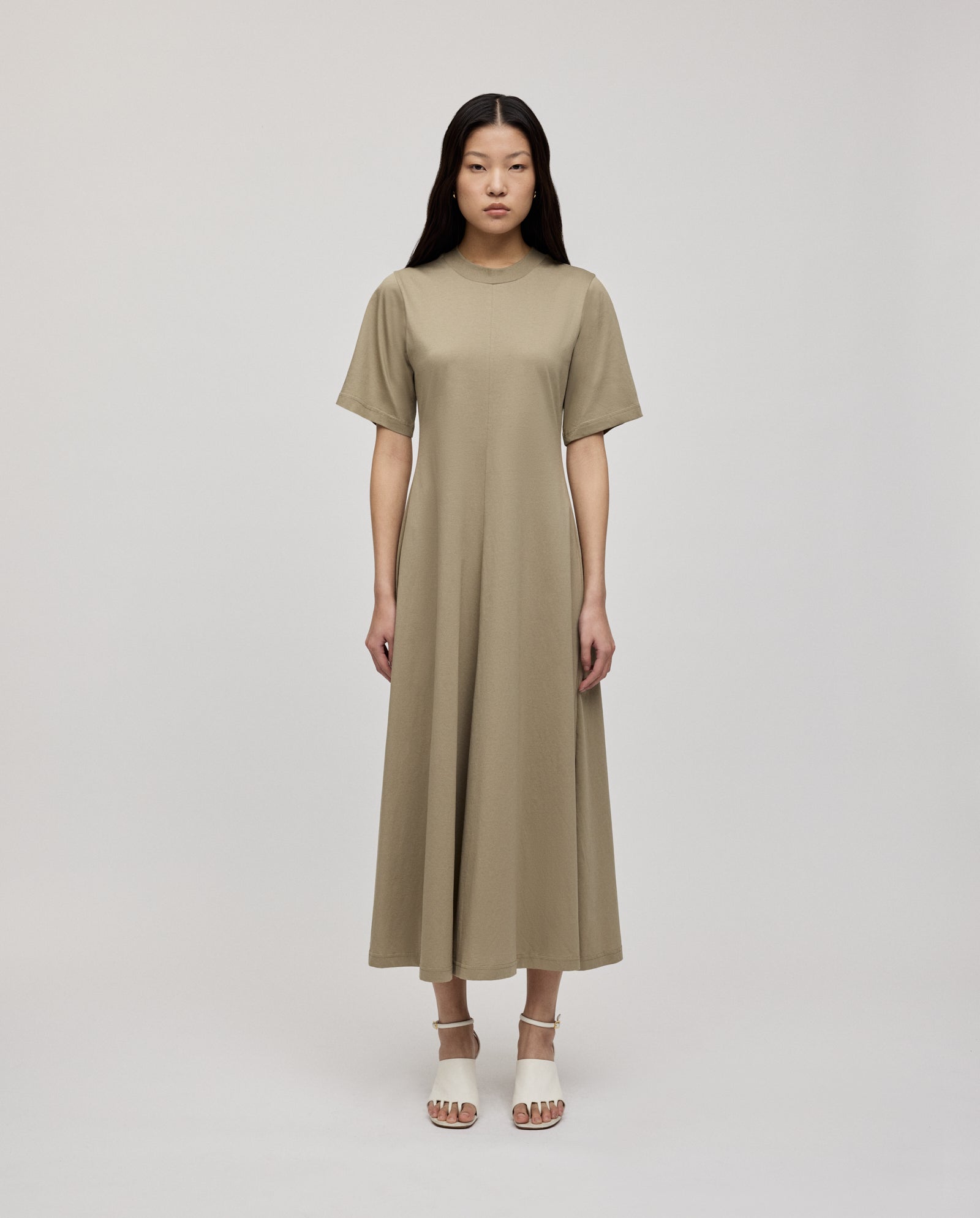 A woman wears the IVY OAK FRANZISKA Jersey Dress in beige—ankle-length, fitted silhouette, short sleeves—paired with white heeled sandals against a plain background.