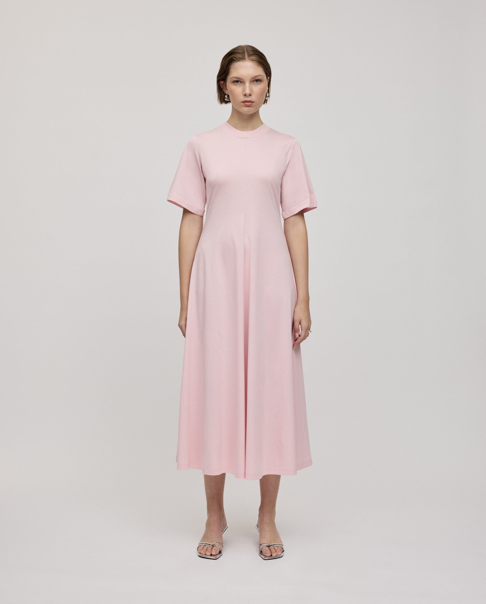 A woman stands facing forward wearing the IVY OAK FRANZISKA Jersey Dress, a light pink, short-sleeve A-line style with an ankle-length fitted silhouette and sandals.