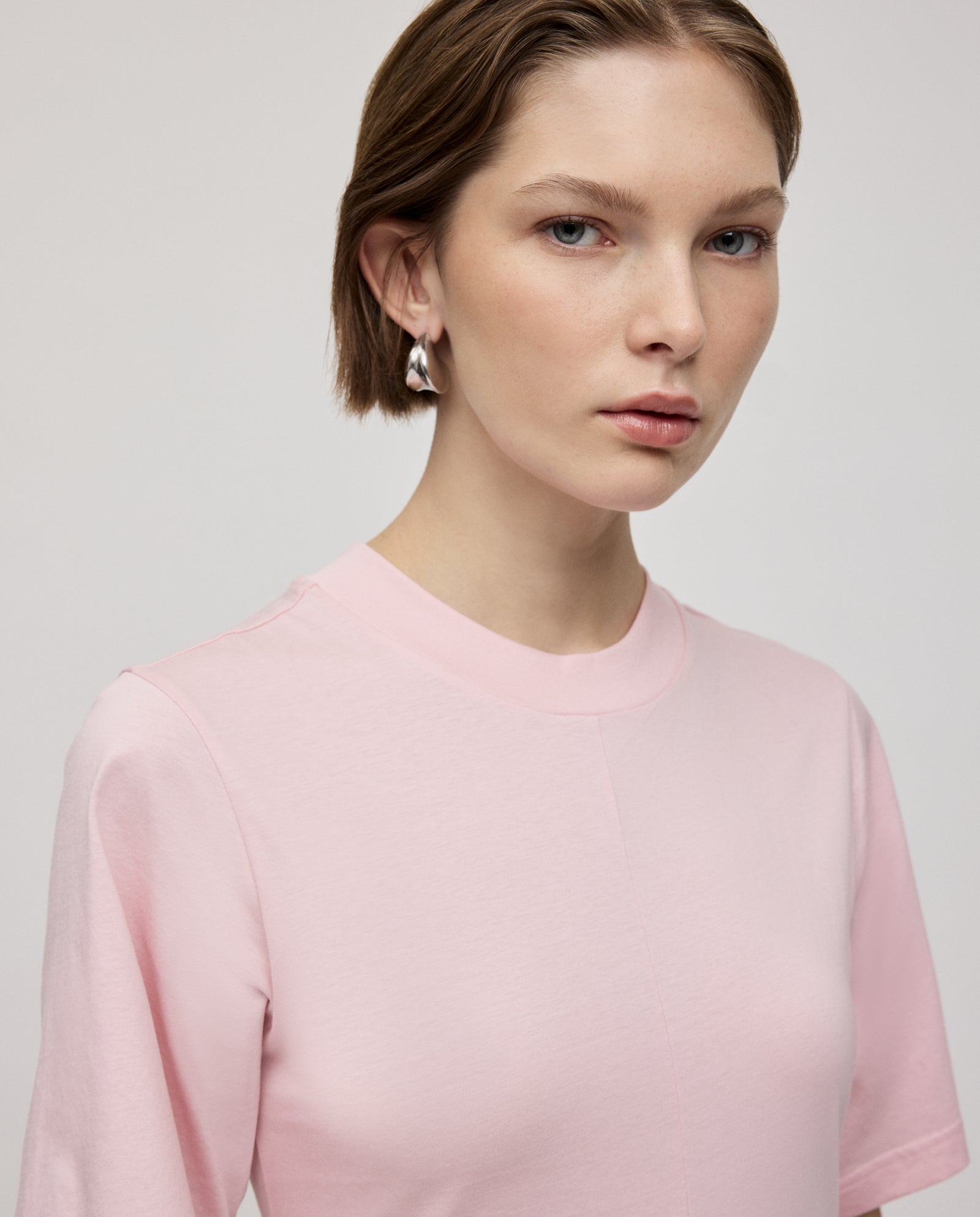 Woman with short brown hair wears the IVY OAK FRANZISKA Jersey Dress in light pink and pearl earrings, looking at the camera.