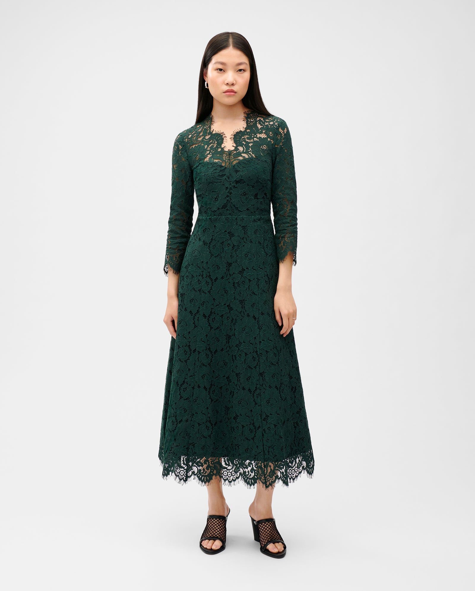 A woman models the IVY OAK MADELEINE Dress in dark green with a long V-neck and stylish black heels, standing against a plain backdrop.