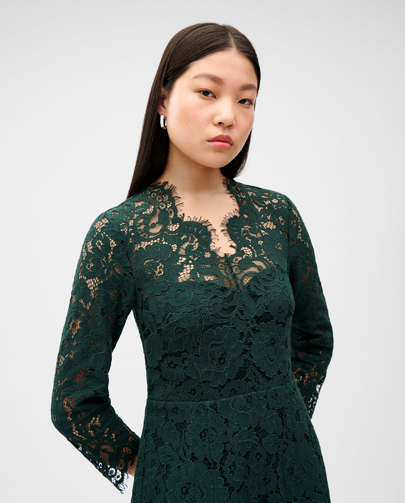 A woman wears the IVY OAK MADELEINE Dress, a dark green midi lace dress with long sleeves and an elegant back cutout, standing against a plain light background.