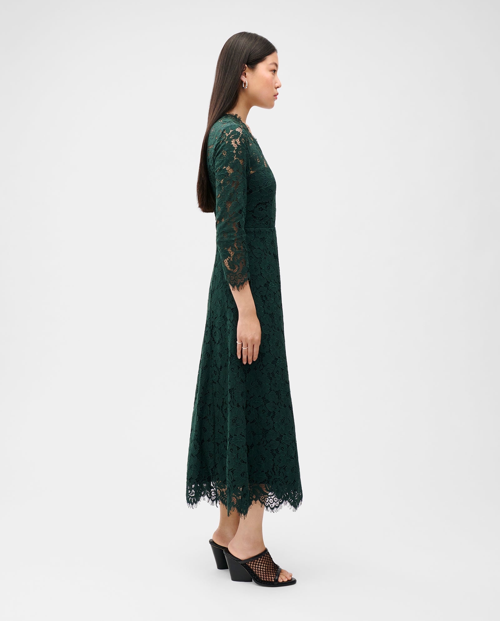 Frau trägt das dunkelgrüne MADELEINE Midi Spitzenkleid von IVY OAK mit Rückenausschnitt und schwarzen Heels, steht seitlich vor weißem Hintergrund.