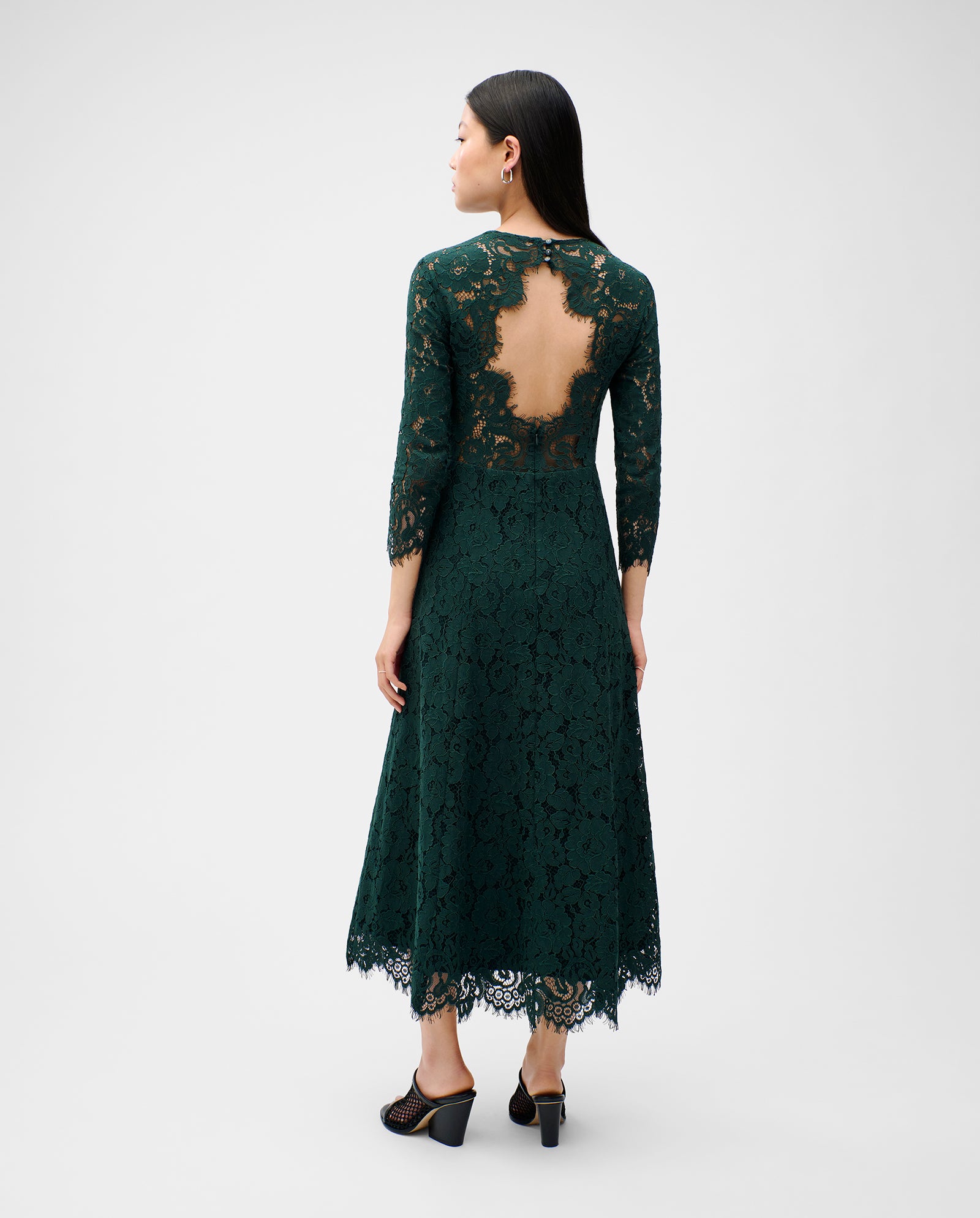 Woman in an elegant dark green MADELEINE Midi dress by IVY OAK, featuring a V-neckline and sheer back detail, standing and facing away.