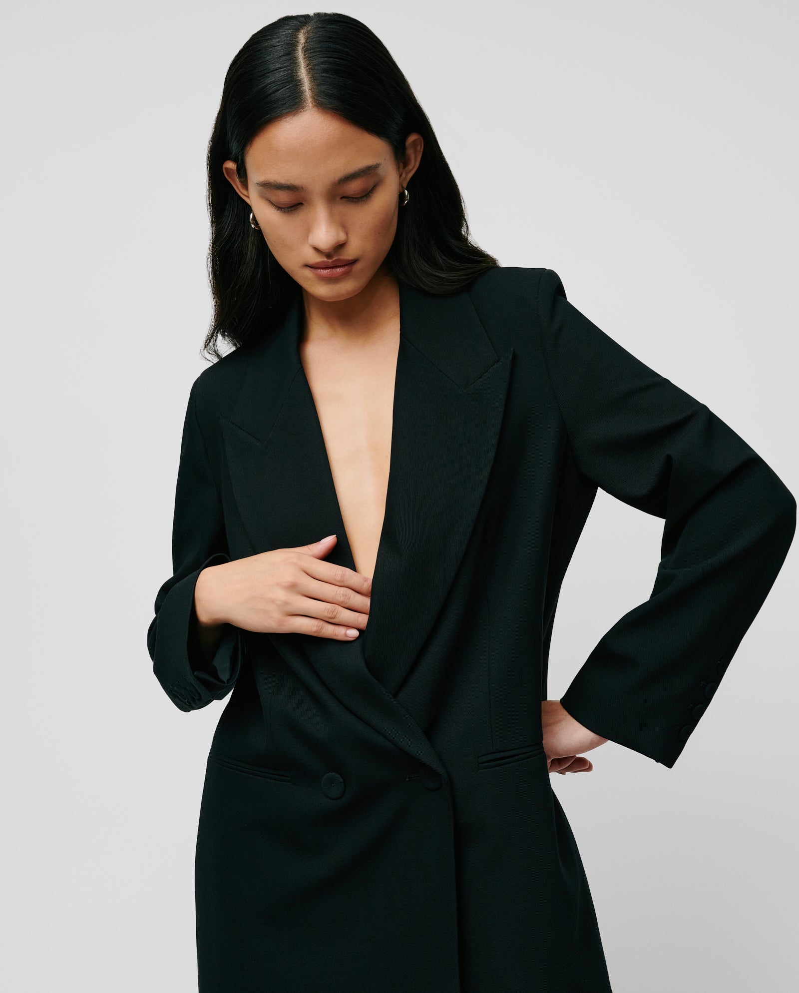 A woman wearing the IVY OAK JIL Blazer in black stands against a plain background, looking down as she adjusts the double-breasted front.