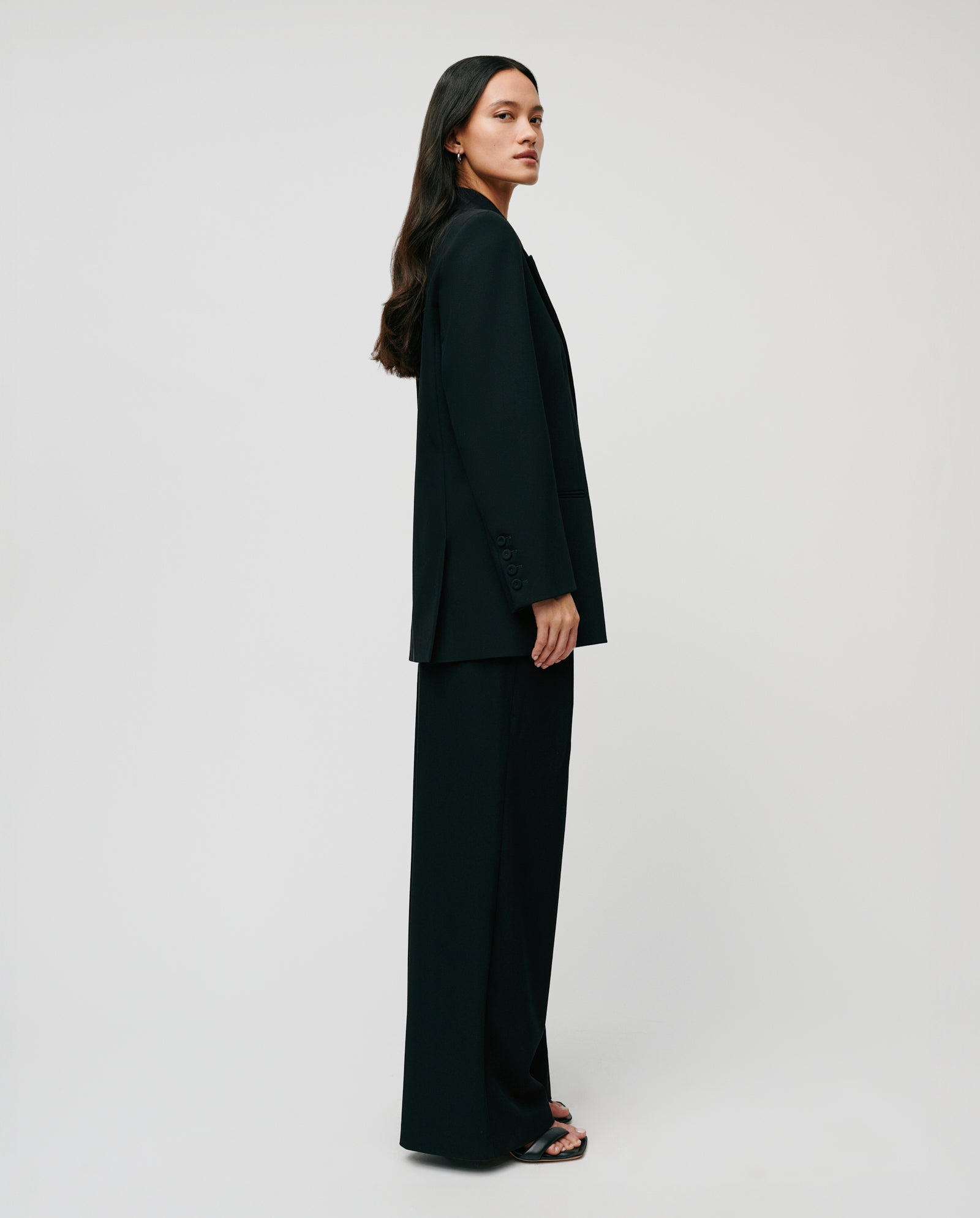 A woman with long dark hair, wearing the IVY OAK JIL Blazer with peak lapels and sandals, stands sideways against a plain light background.