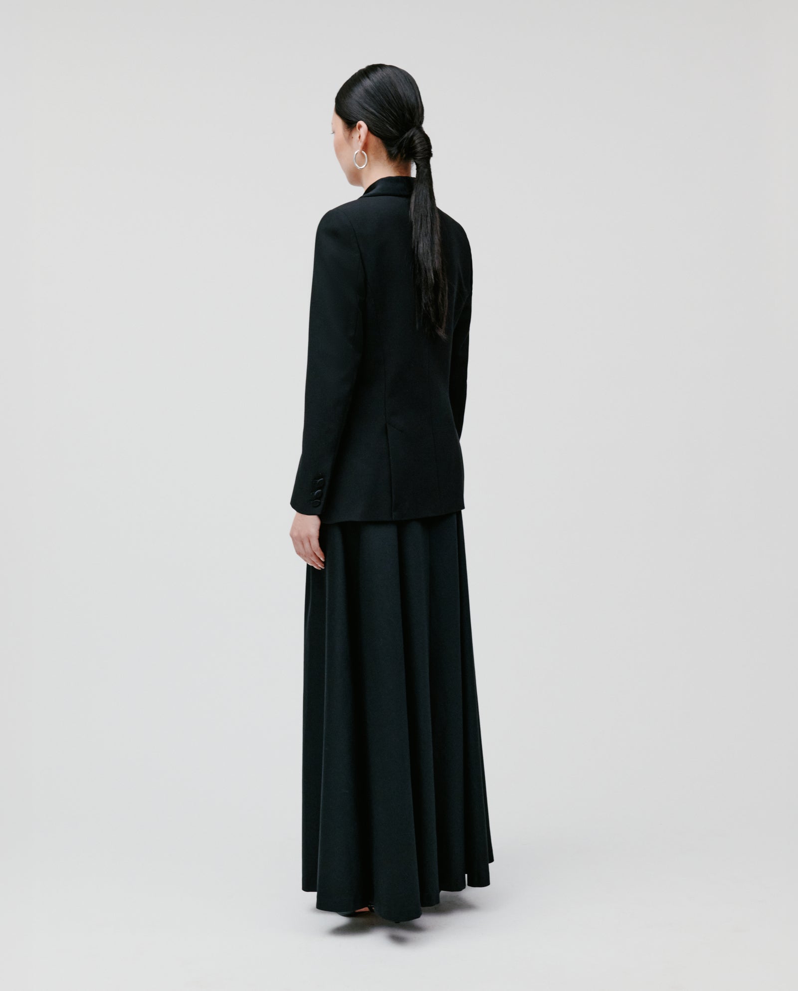 A woman stands facing away against a plain, light background, wearing the IVY OAK JANA Blazer and a long black skirt.