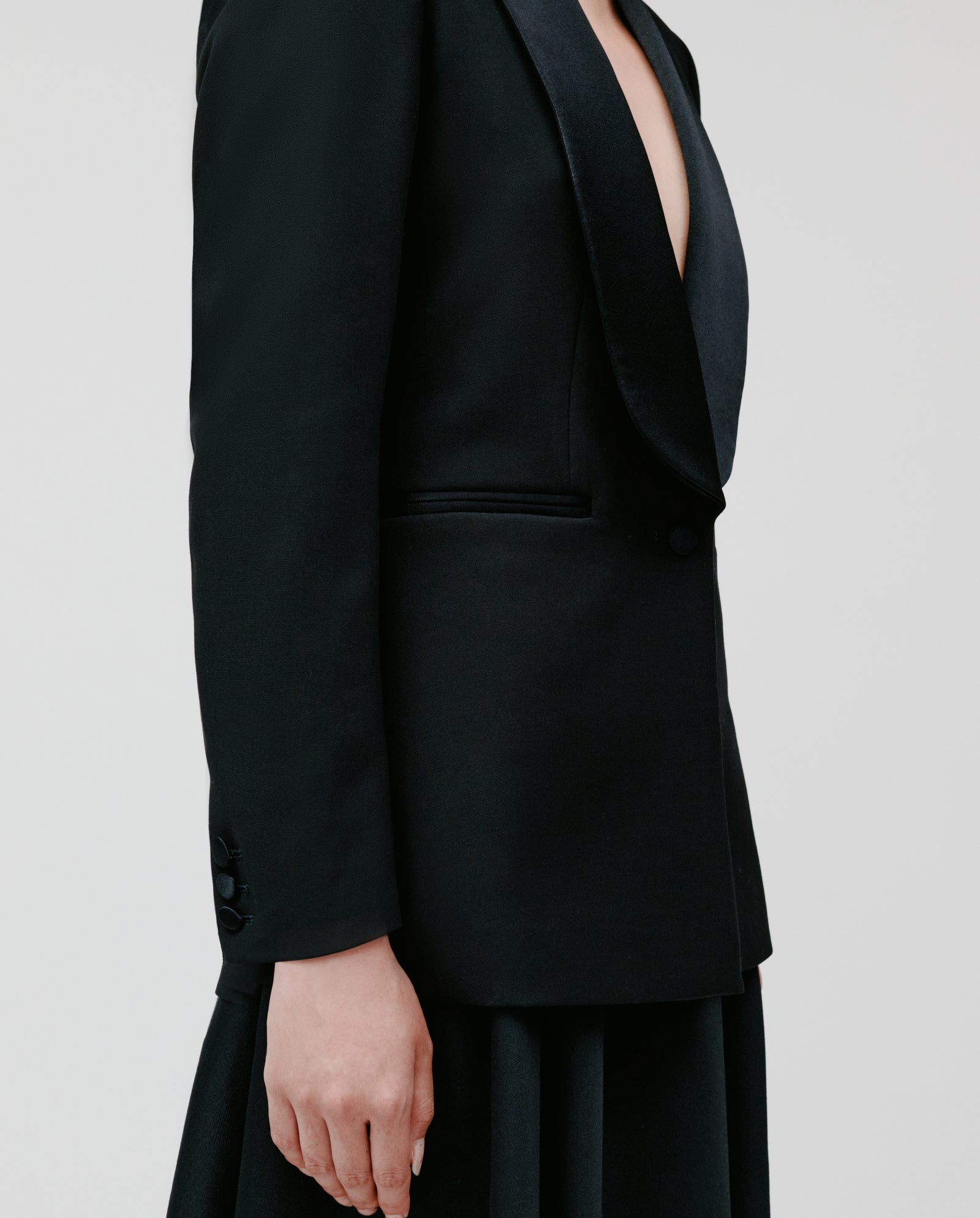 A model wears the JANA Blazer by IVY OAK, featuring a black lined design with a shawl collar, paired with a black skirt and shown in profile against a plain backdrop.