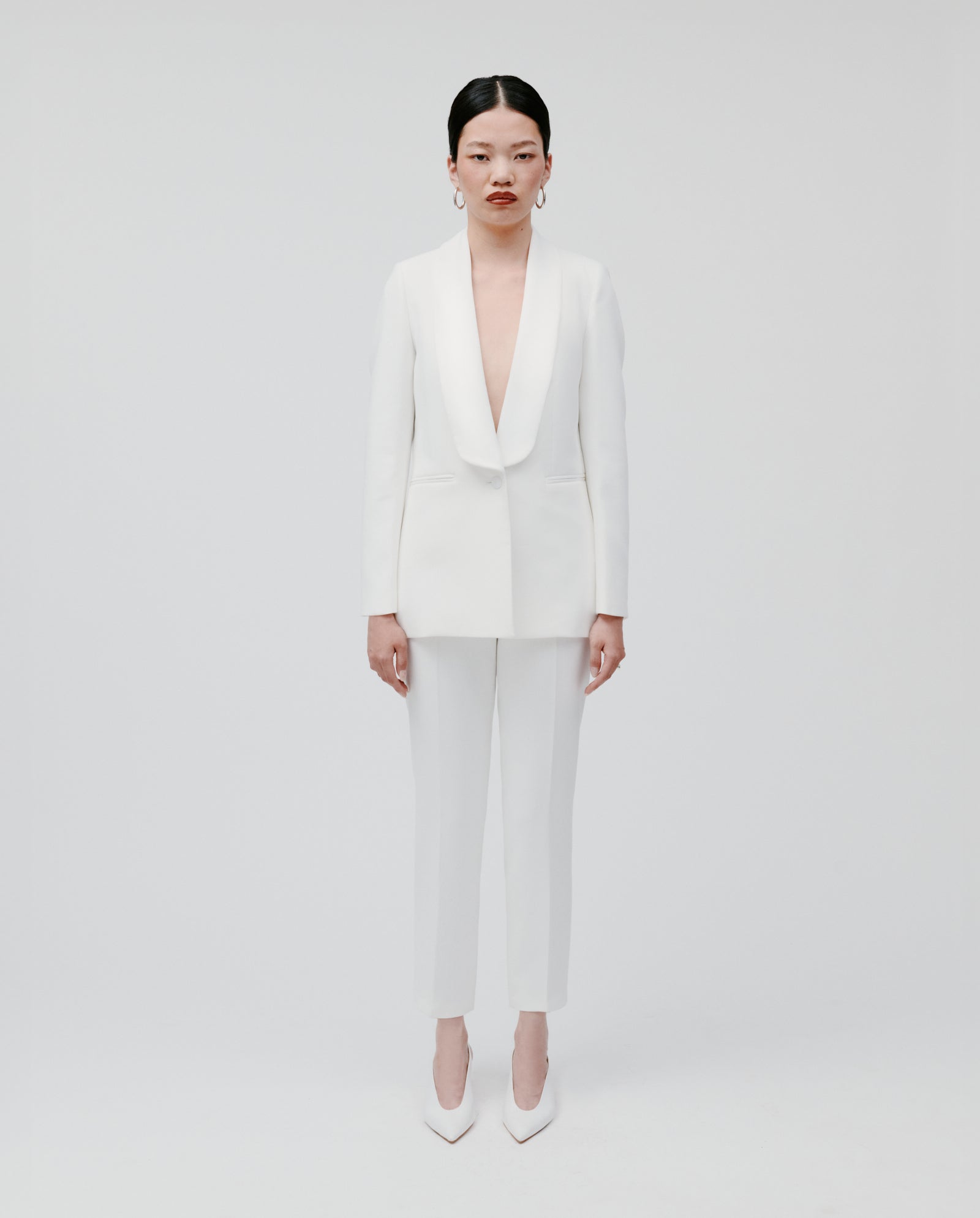 A woman wears the IVY OAK JANA Blazer, a white, lined piece with a sharp shawl collar, paired with heels as she stands against a plain white background, gazing straight ahead.