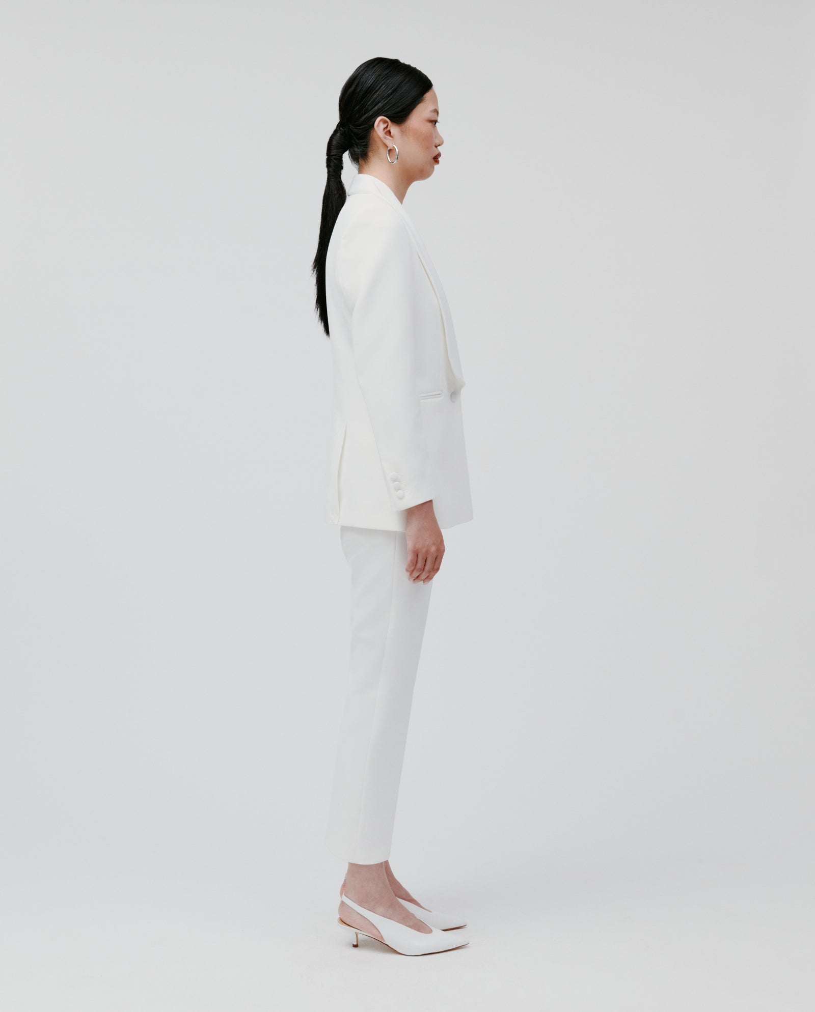 A woman in profile wears the IVY OAK JANA Blazer with a shawl collar and heels, standing against a plain white background.