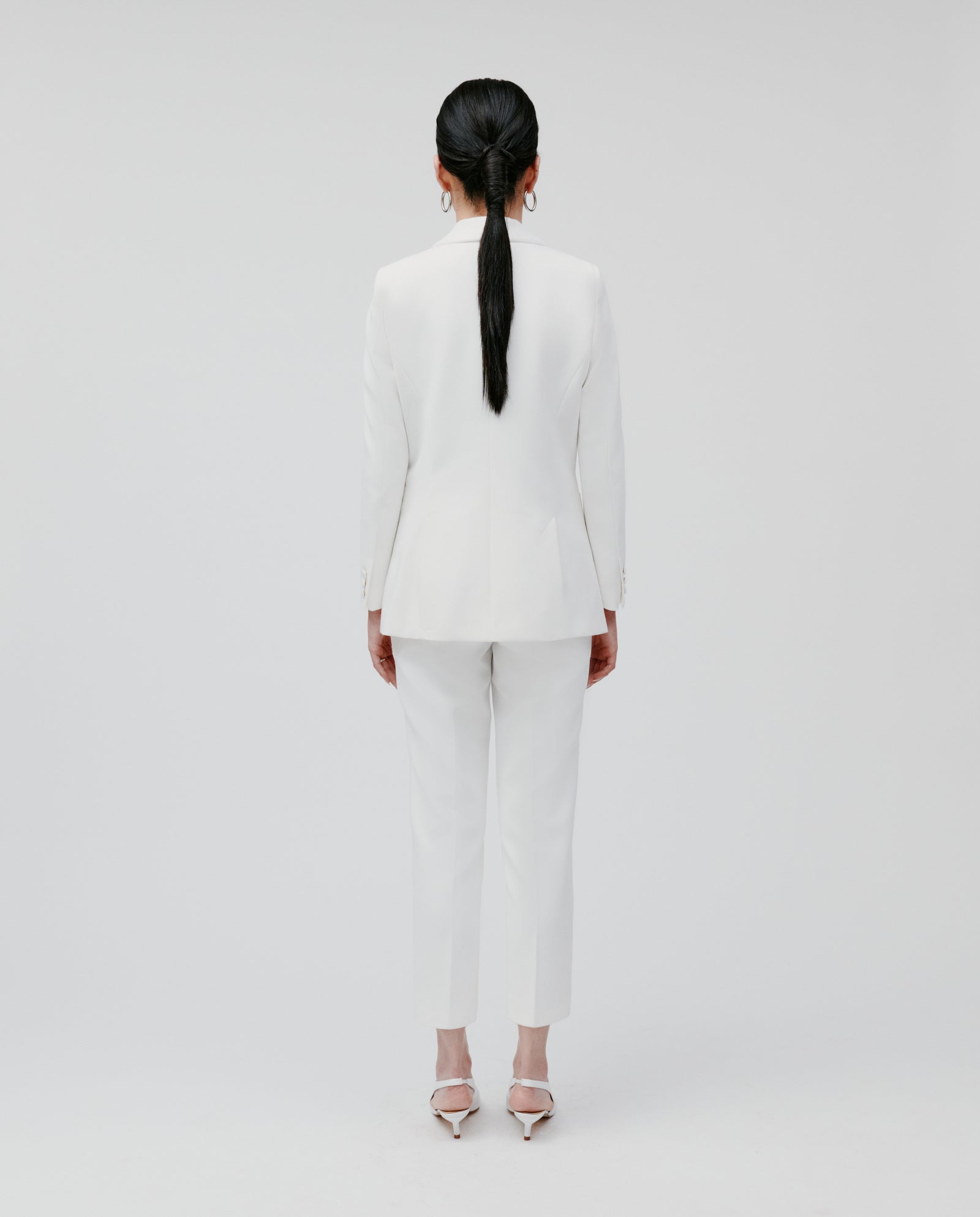 A person stands facing away against a plain white background, wearing the IVY OAK JANA Blazer—white with lining and a shawl collar—paired with heels.