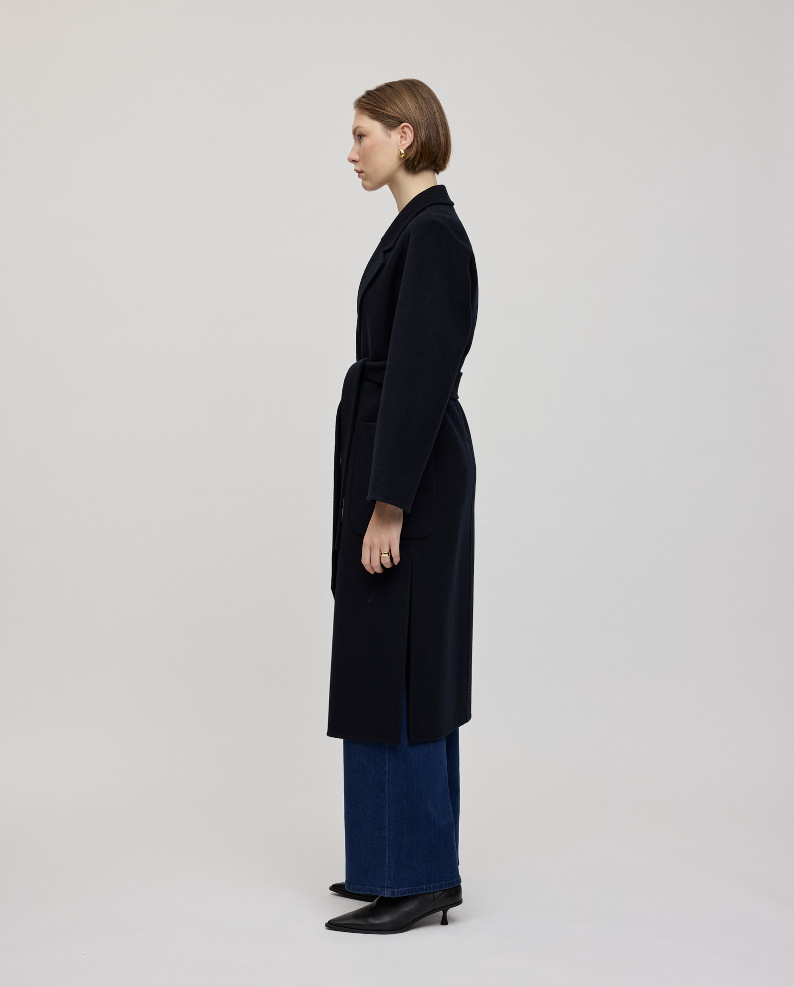 Eine Frau trägt den CELIA Coat von IVY OAK aus Responsible Wool Standard in Midilänge, kombiniert mit blauen Jeans und schwarzen Boots, und steht im Profil vor einem schlichten Hintergrund.