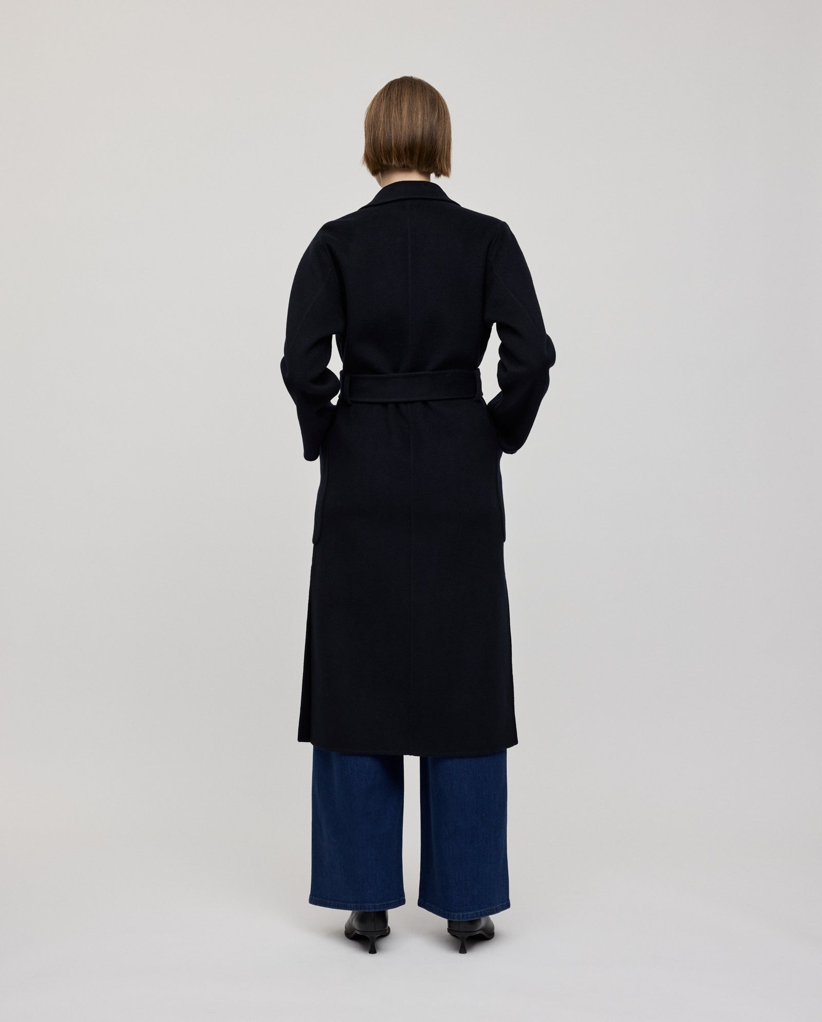 A person faces away, wearing the IVY OAK CELIA Coat in black wool (mid-length), paired with wide-leg jeans and black shoes against a plain background. Made according to the Responsible Wool Standard for ethical fashion.