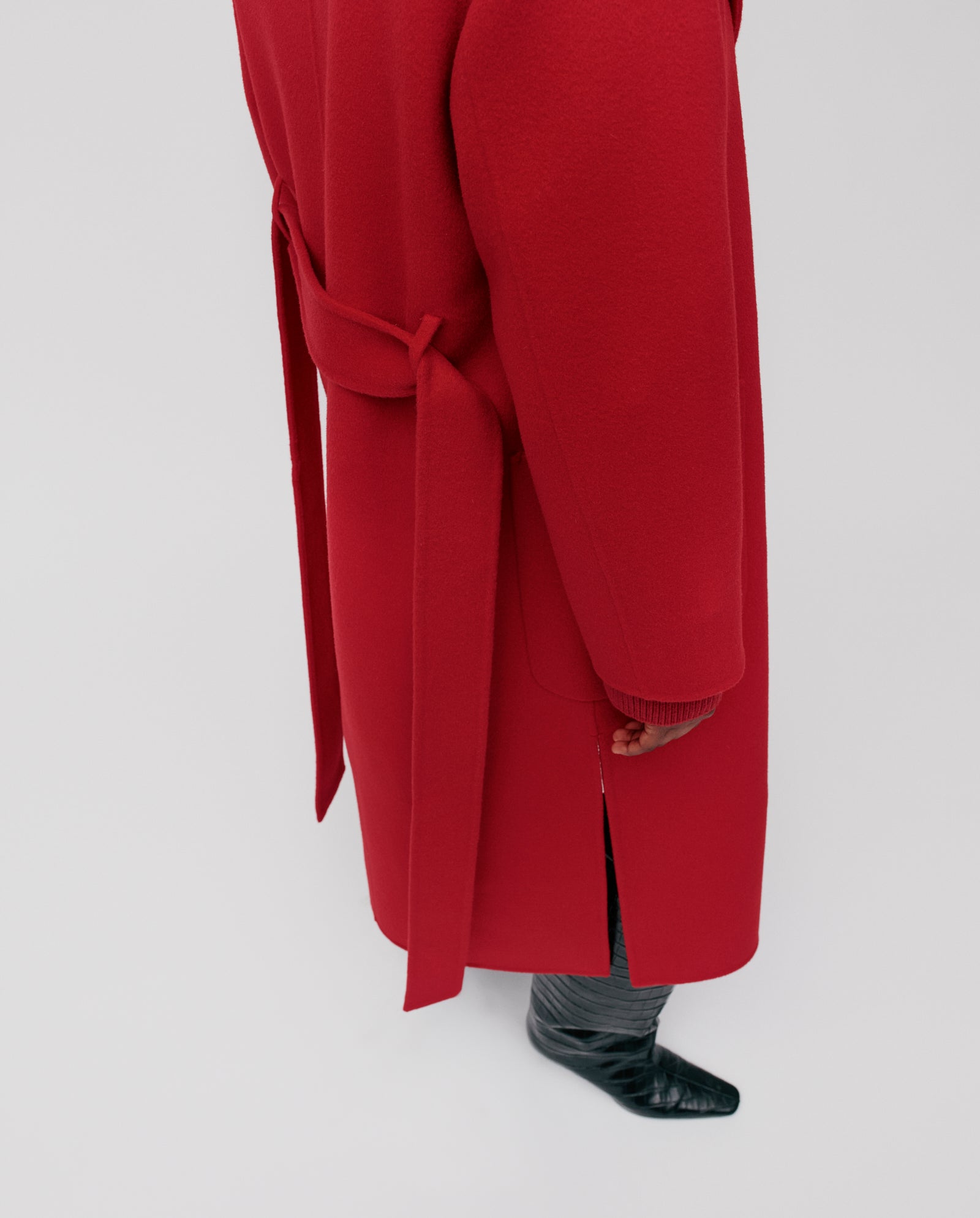 A person wears the IVY OAK CELIA midi red wool coat, belted at the back, with black boots, standing on a light surface.