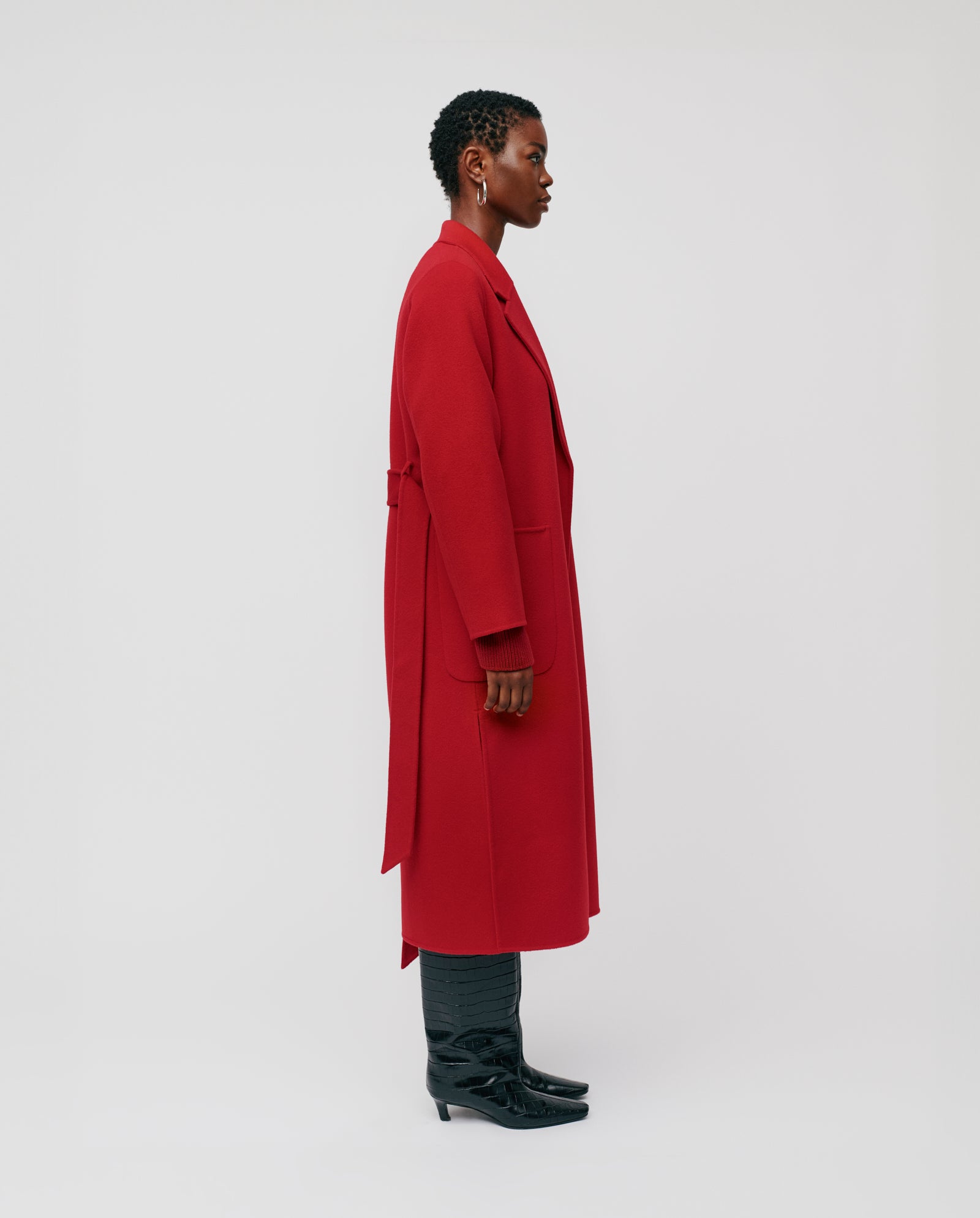 A woman stands in profile against a plain light background wearing the IVY OAK CELIA midi red wool coat, certified by the Responsible Wool Standard, paired with black boots.