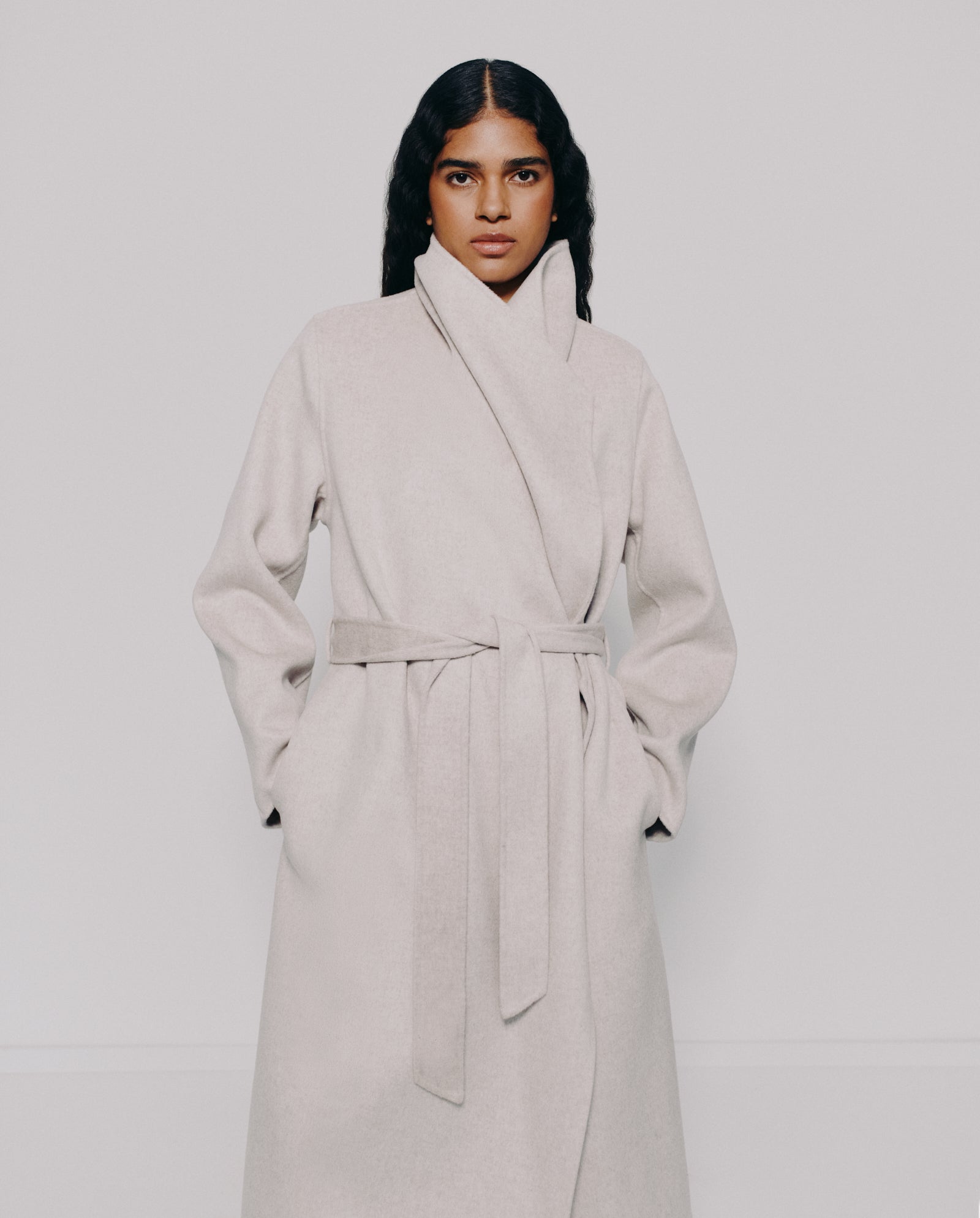 A woman models the IVY OAK CARRIE ROSE Coat, a minimalistic long light beige wool design featuring a high collar and belt, set against a plain background.