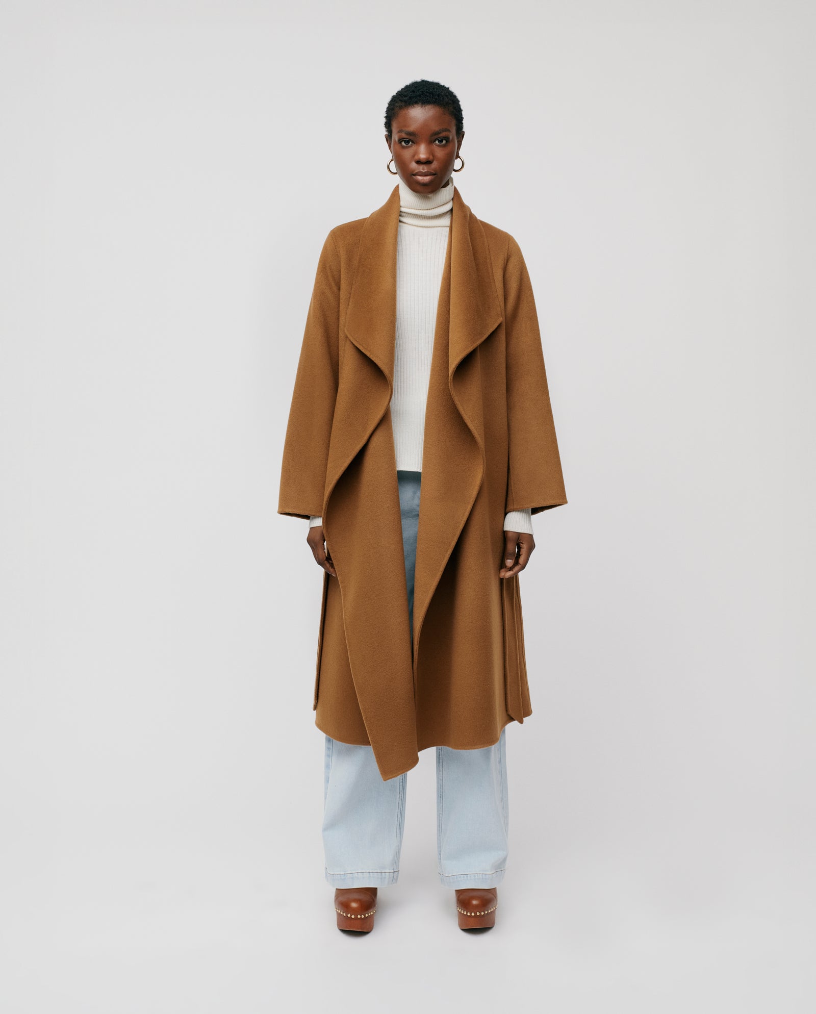 Woman wearing the IVY OAK CARRIE ROSE Coat, featuring a big lapel collar, made from Responsible Wool Standard certified materials, styled with a white turtleneck, light jeans, and brown platform shoes against a plain background.