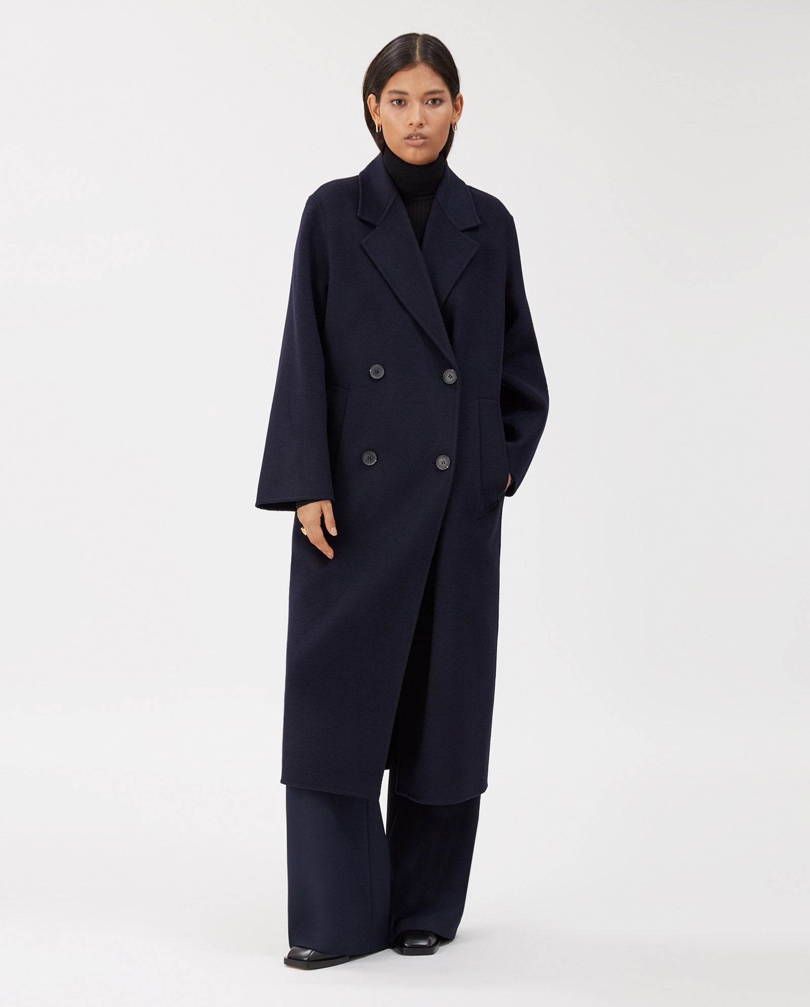 A woman wears the IVY OAK CLARA Coat—an oversized, dark navy midi wool coat made to Responsible Wool Standard—styled over wide-leg pants and a black turtleneck.