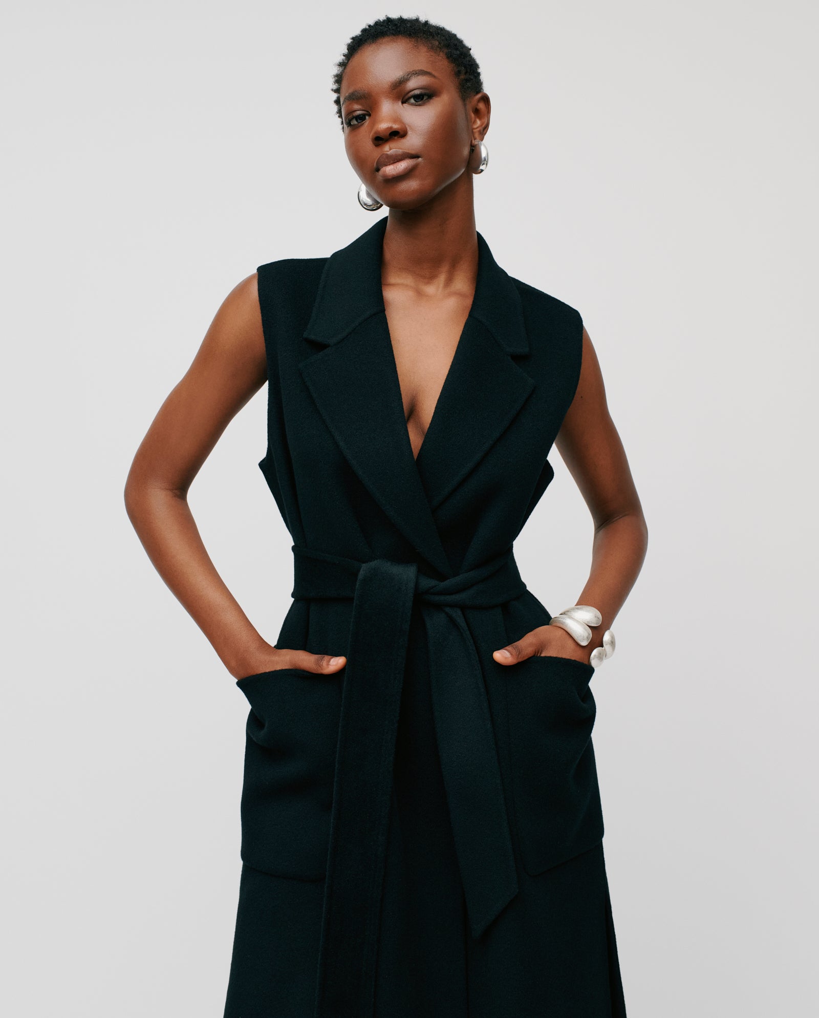 A woman wears a sleeveless black CLARISSA Vest by IVY OAK (DE), made from RWS wool, and poses with her hands in the pockets against a plain background.