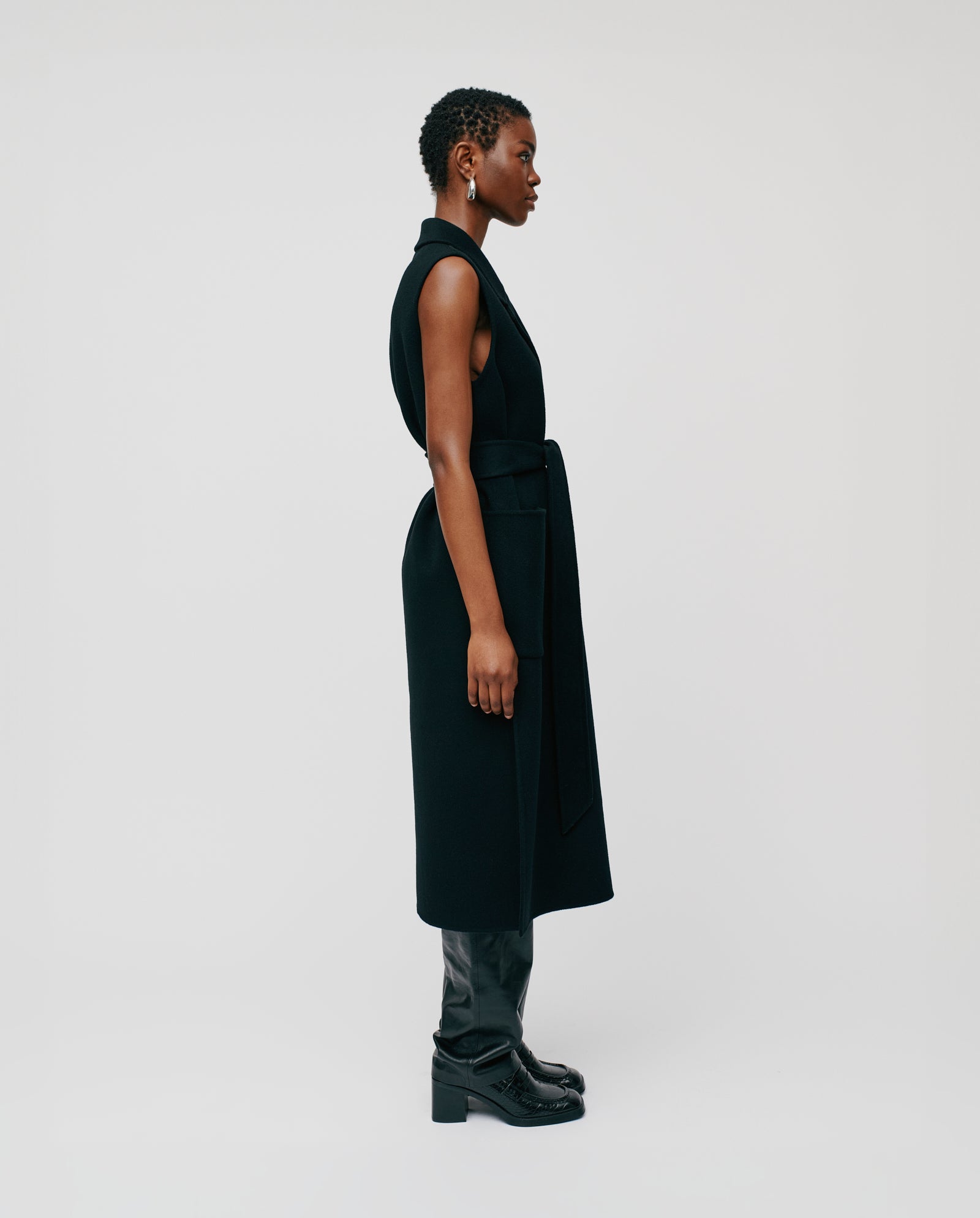 A woman stands in profile against a plain background wearing the sleeveless black CLARISSA Vest by IVY OAK (DE), crafted from RWS wool, paired with black pants and boots.
