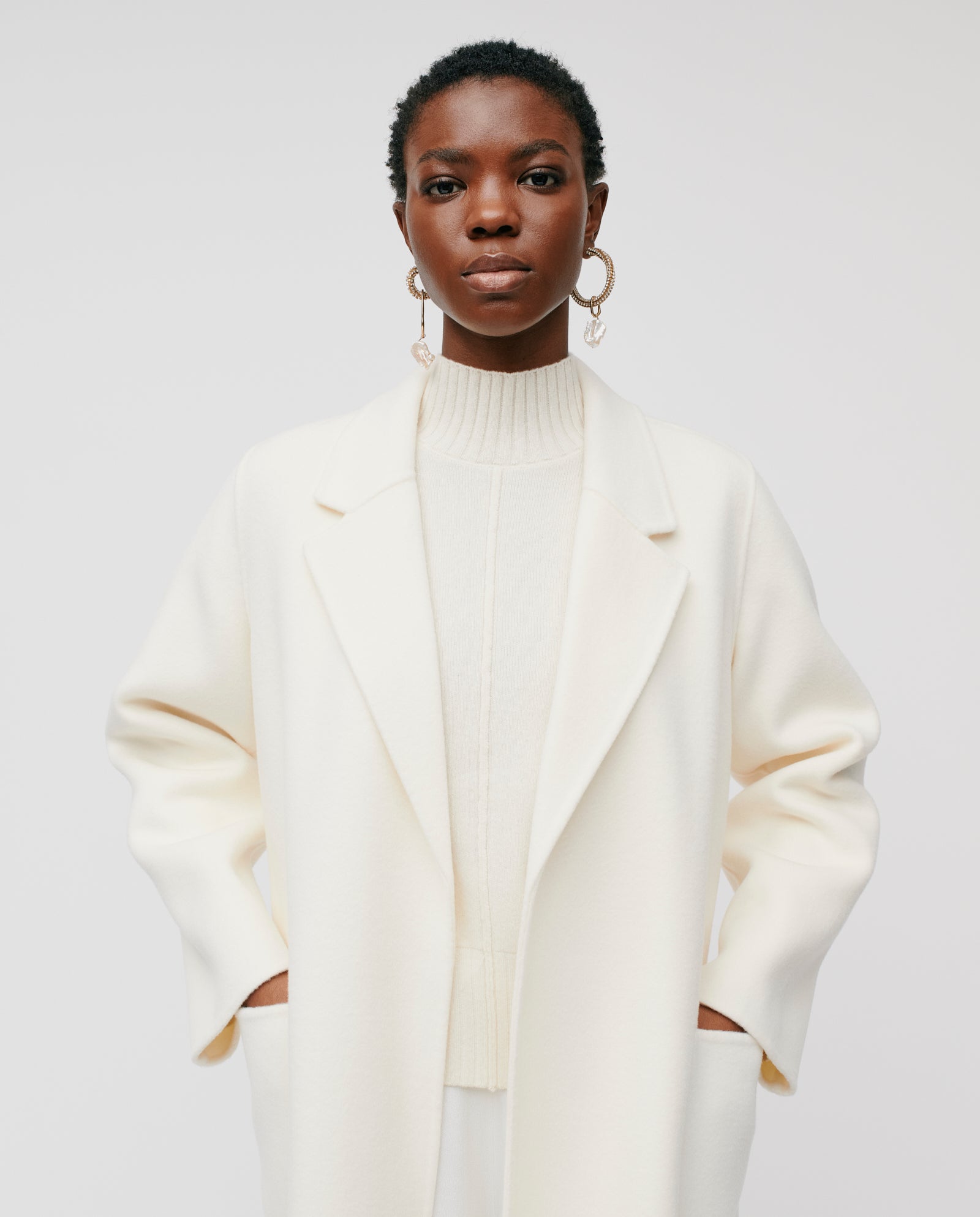 A woman wears the IVY OAK CELIA Wool Coat in cream, featuring a loose fit and turtleneck, with hands in pockets against a plain white background.