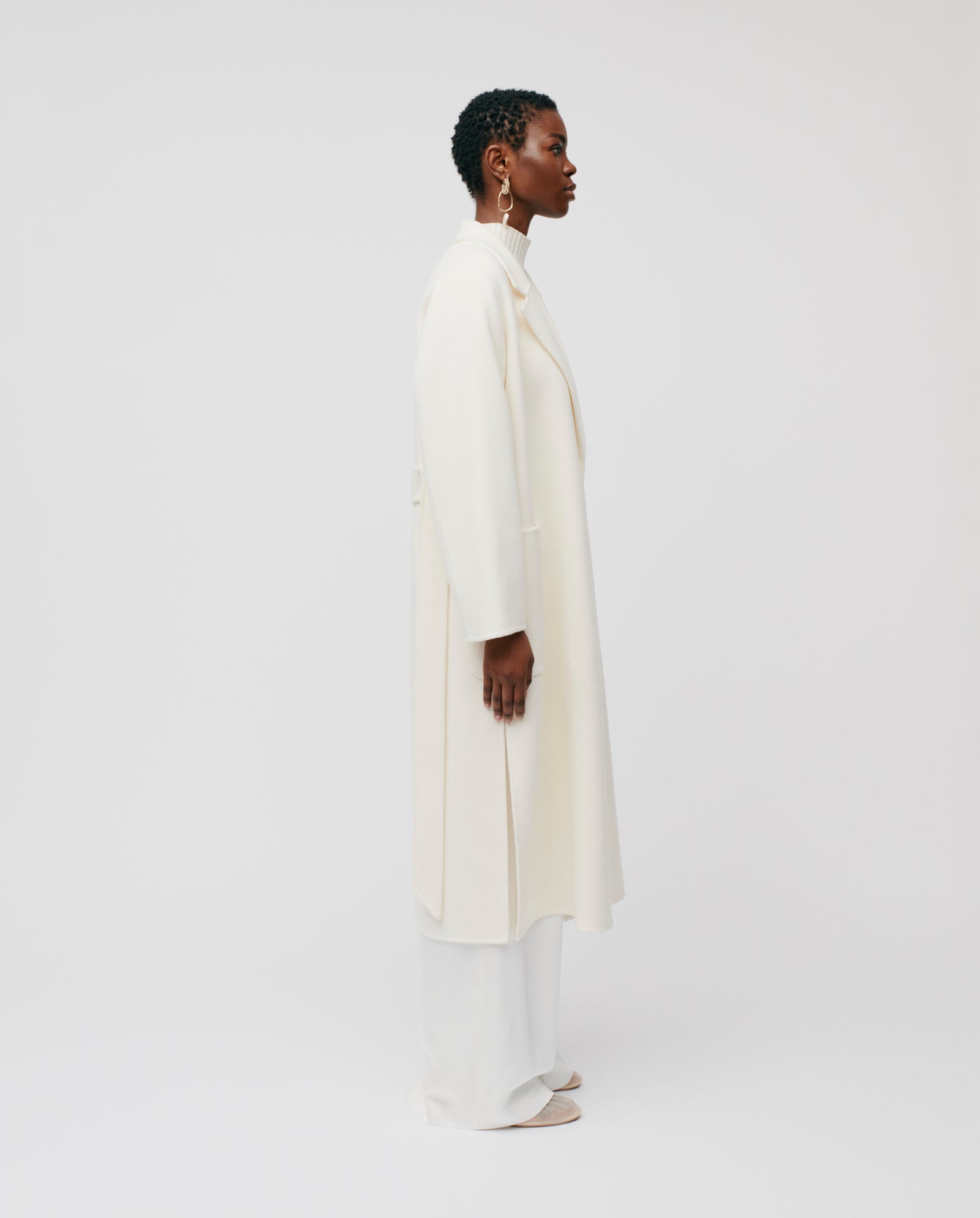 A woman in profile wears the IVY OAK CELIA Wool Coat in a loose fit, paired with wide-leg white pants and earrings, set against a white background.