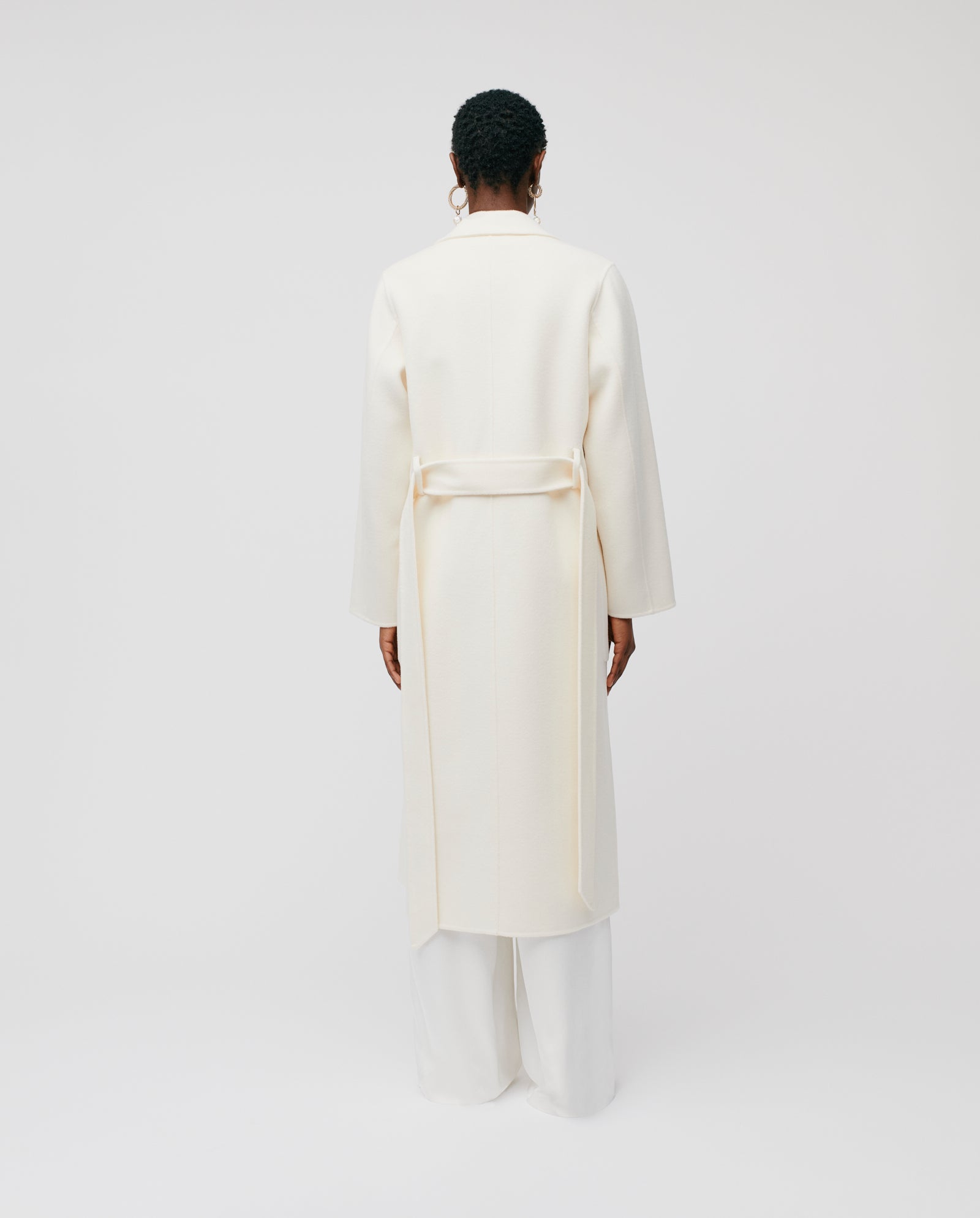 A person stands with their back to the camera, wearing the IVY OAK CELIA Wool Coat in a loose fit midi length, paired with white pants against a plain background.