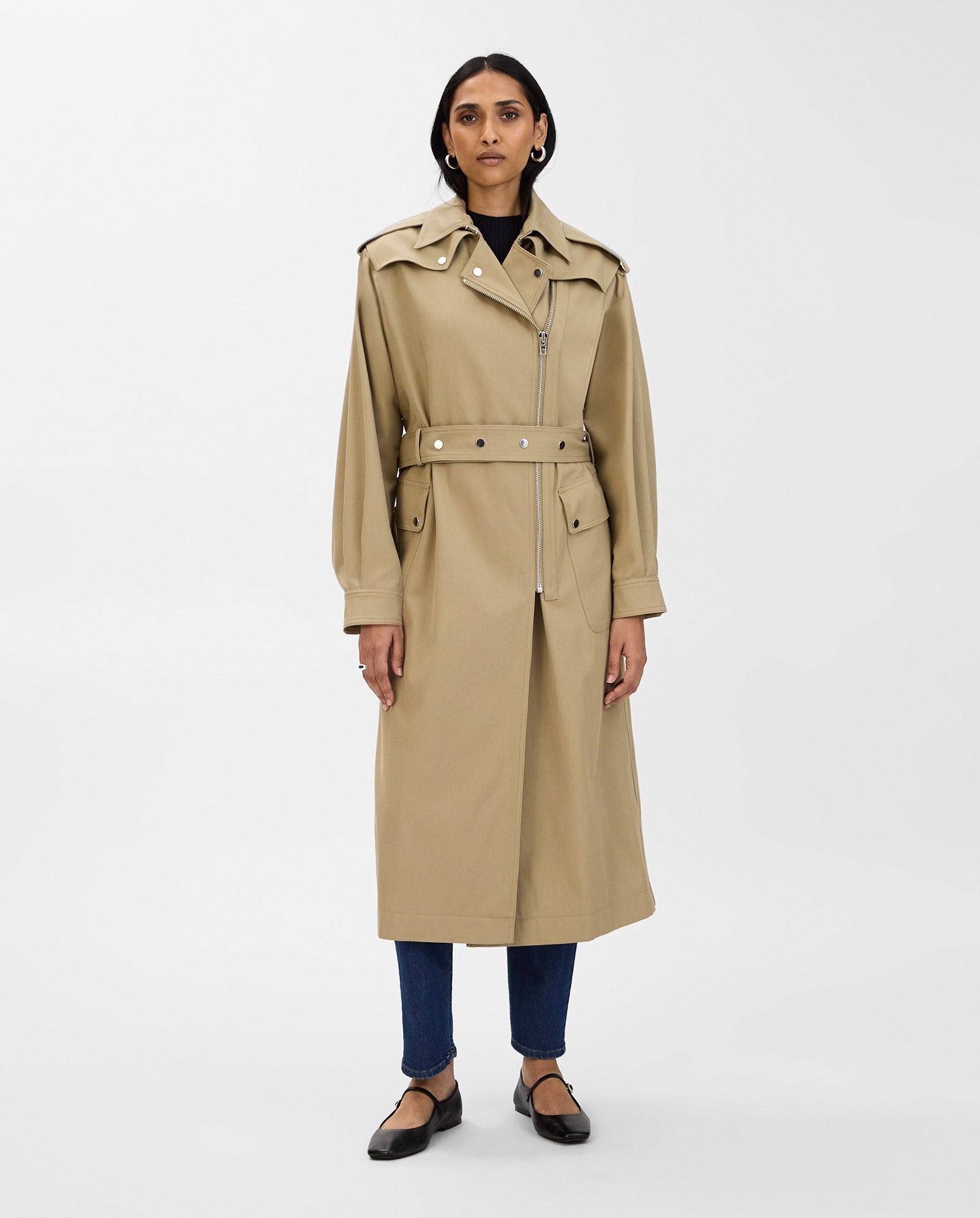 A woman wears the IVY OAK CIARA Coat, an oversized beige trenchcoat with an adjustable belt, styled with dark pants and black flats against a plain background.