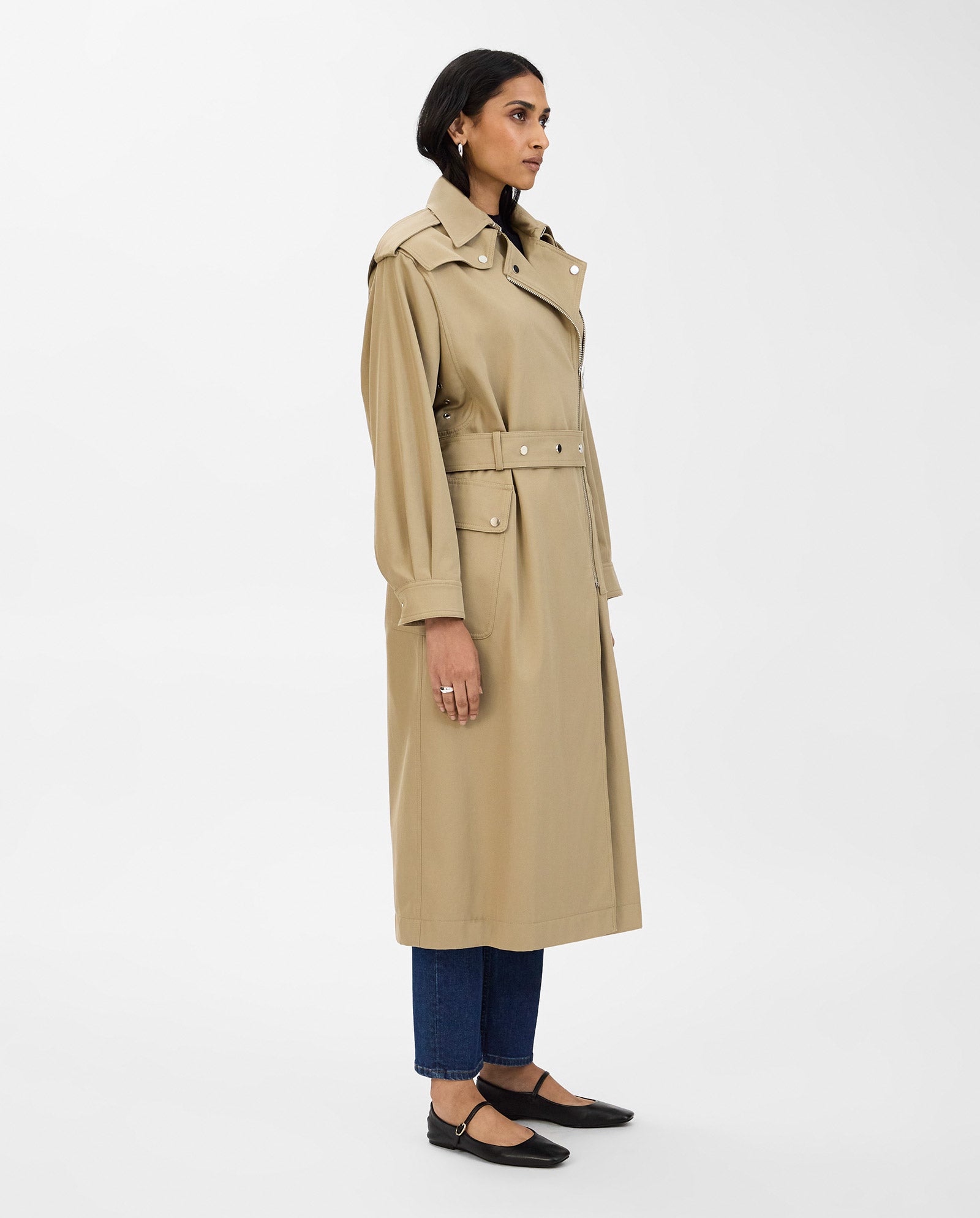 A woman stands sideways against a plain white background wearing the IVY OAK CIARA Coat in an oversized beige style with an adjustable belt, paired with blue jeans and black flats.