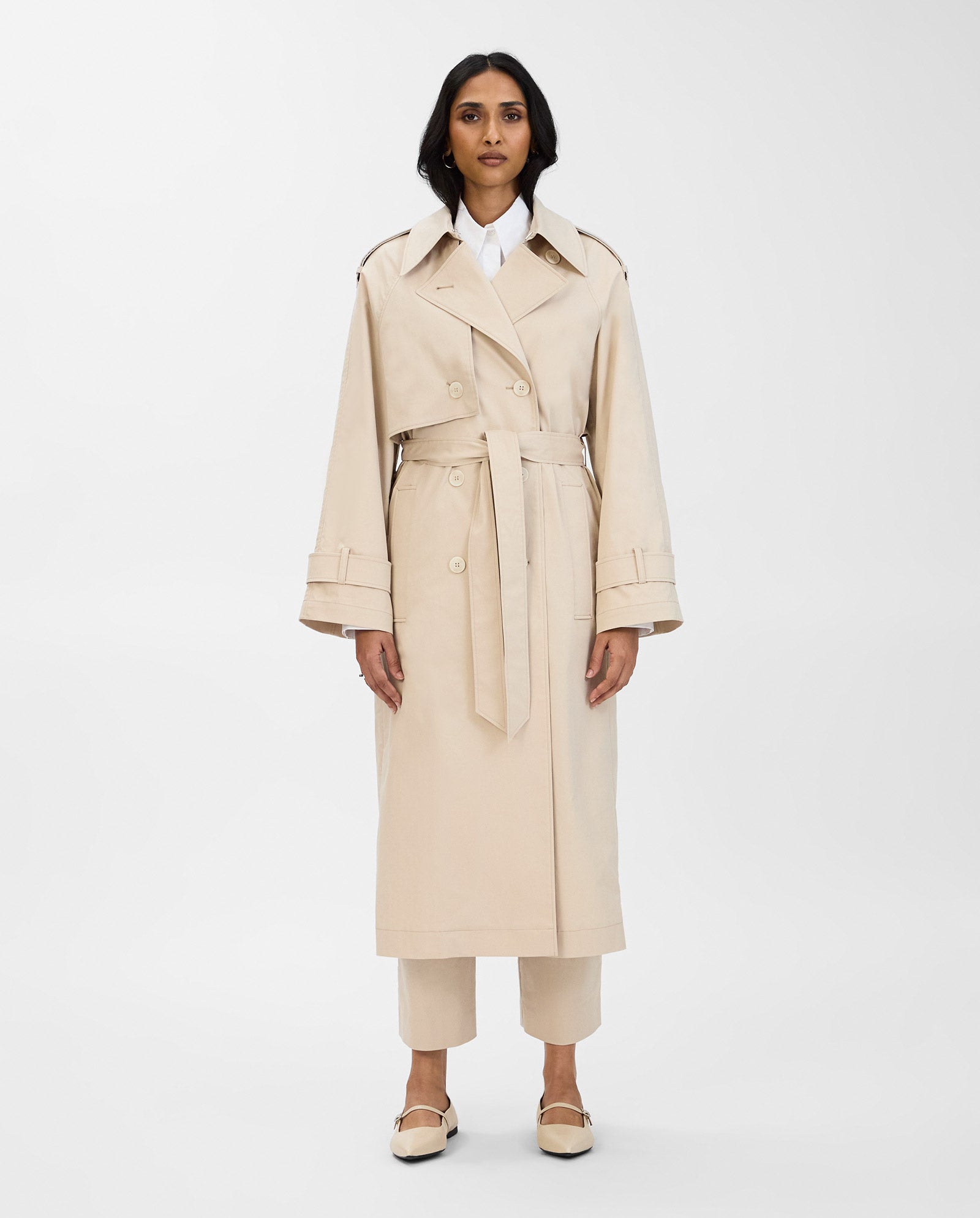 A woman stands against a plain background wearing the IVY OAK CORALINE Coat, a classic double-breasted trenchcoat crafted from organic cotton, paired with a white shirt, beige pants, and cream shoes.