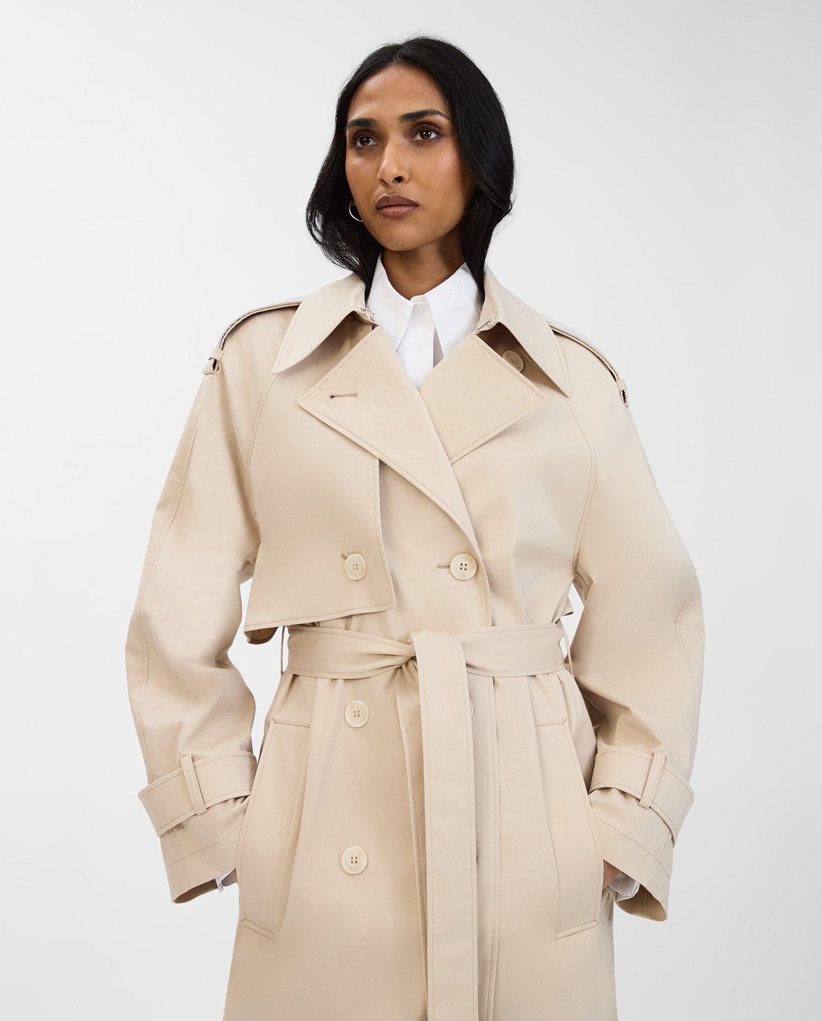 A woman wears the IVY OAK CORALINE Coat, a classic double-breasted trench with belt, made from organic cotton, standing against a plain white background.