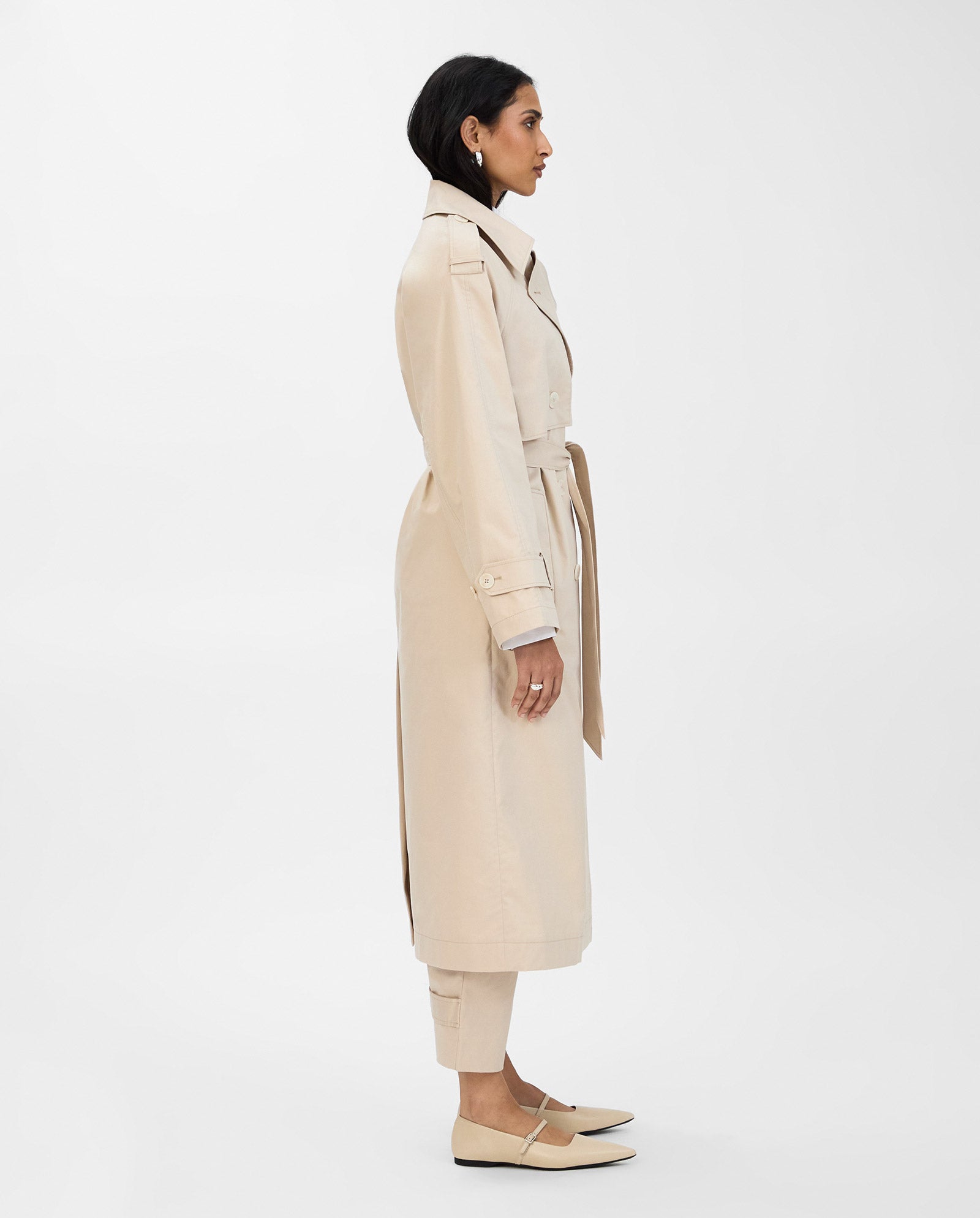 A woman in the IVY OAK CORALINE Coat, a classic double-breasted trench crafted from organic cotton, paired with matching pants, stands in profile against a plain white background.