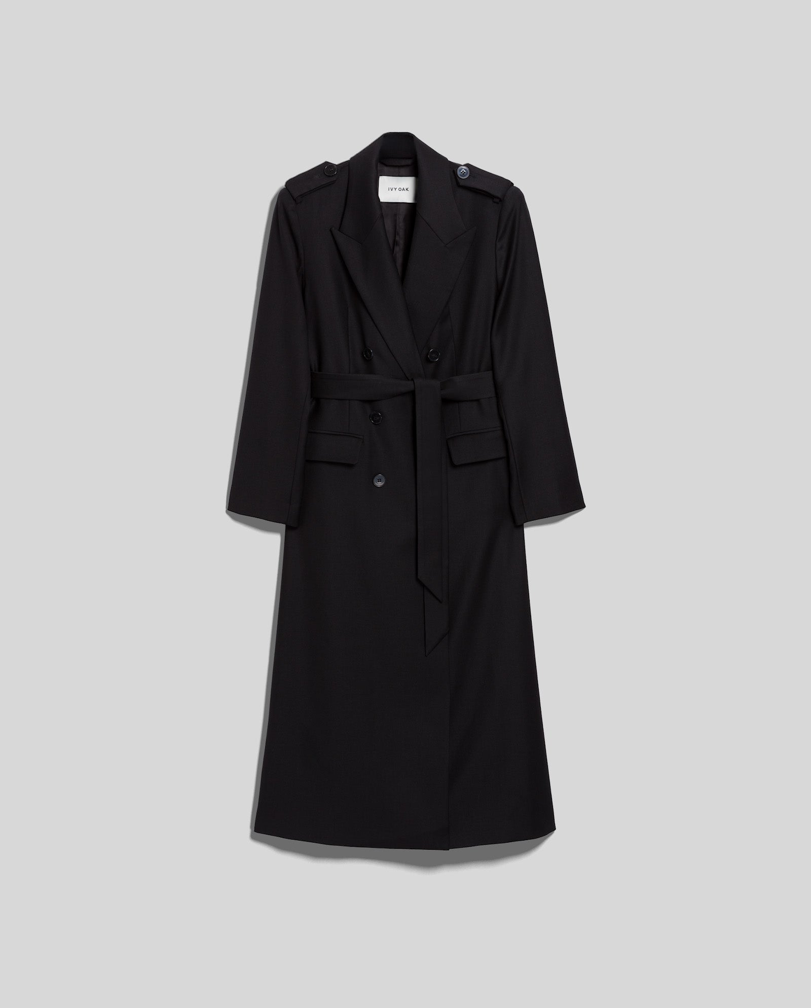 The IVY OAK COLETTE Coat is a long black trench coat featuring a belt, button details, and wide lapels, shown against a plain grey background.