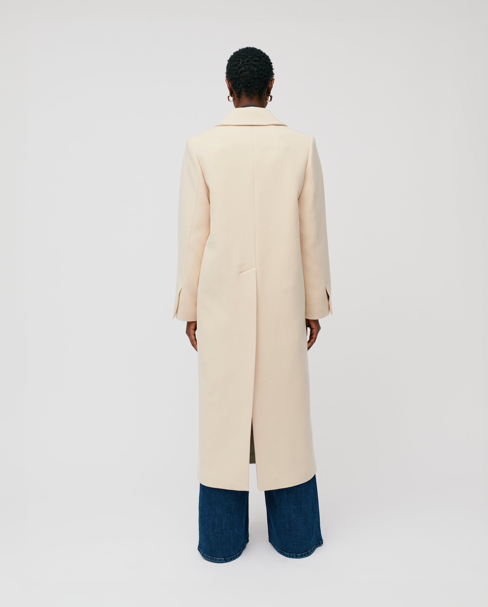 Model wears the IVY OAK CLEMENCE Coat in light beige, styled with wide-leg blue jeans, facing away against a plain background.